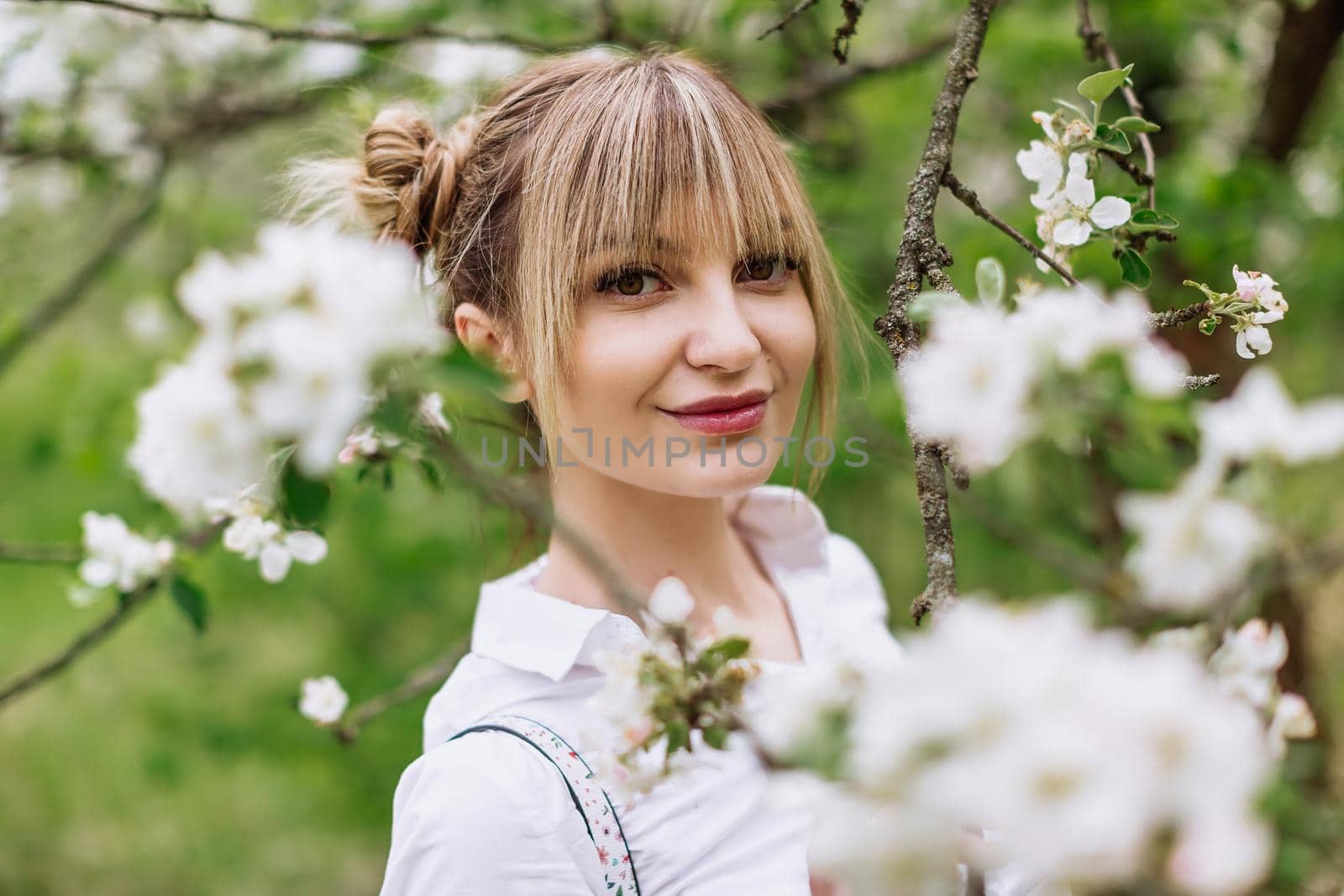 Close-up portrait of beautiful young blonde woman in white shirt with backpack posing under apple tree in blossom in Spring garden by OnPhotoUa