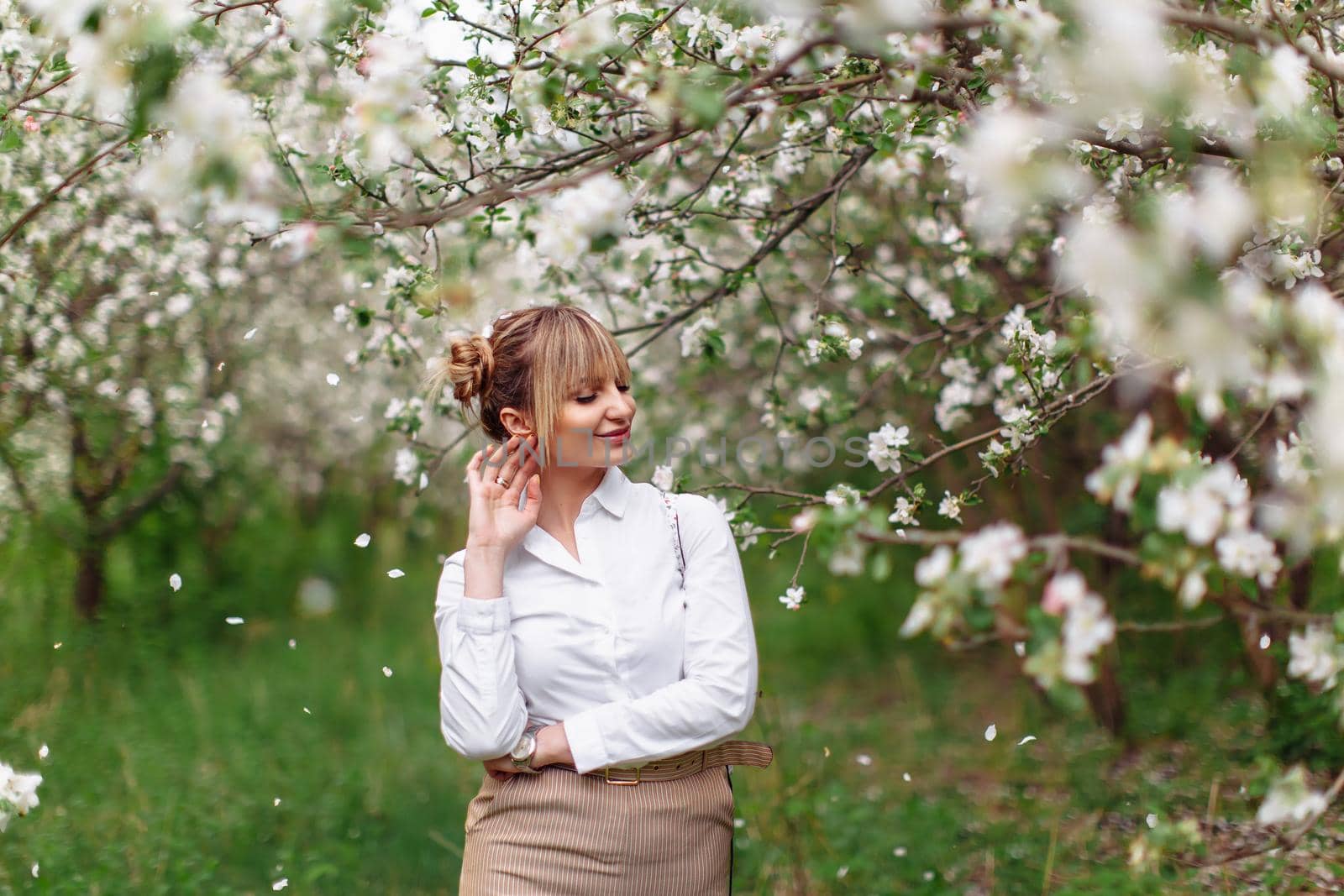 Beautiful young blonde woman in white shirt posing under apple tree in blossom in Spring garden by OnPhotoUa