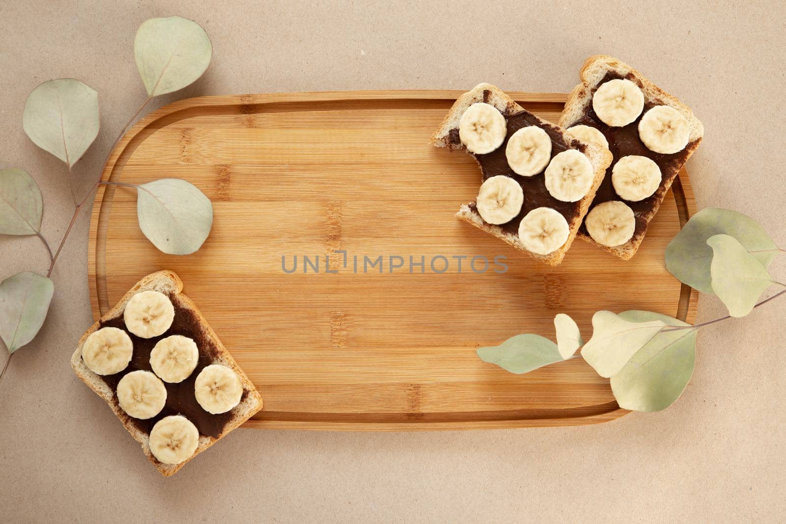 Three banana white bread toasts smeared with chocolate butter that lie on a cutting board with a sprig of leaves on craft paper background. top view with area for text by lunarts