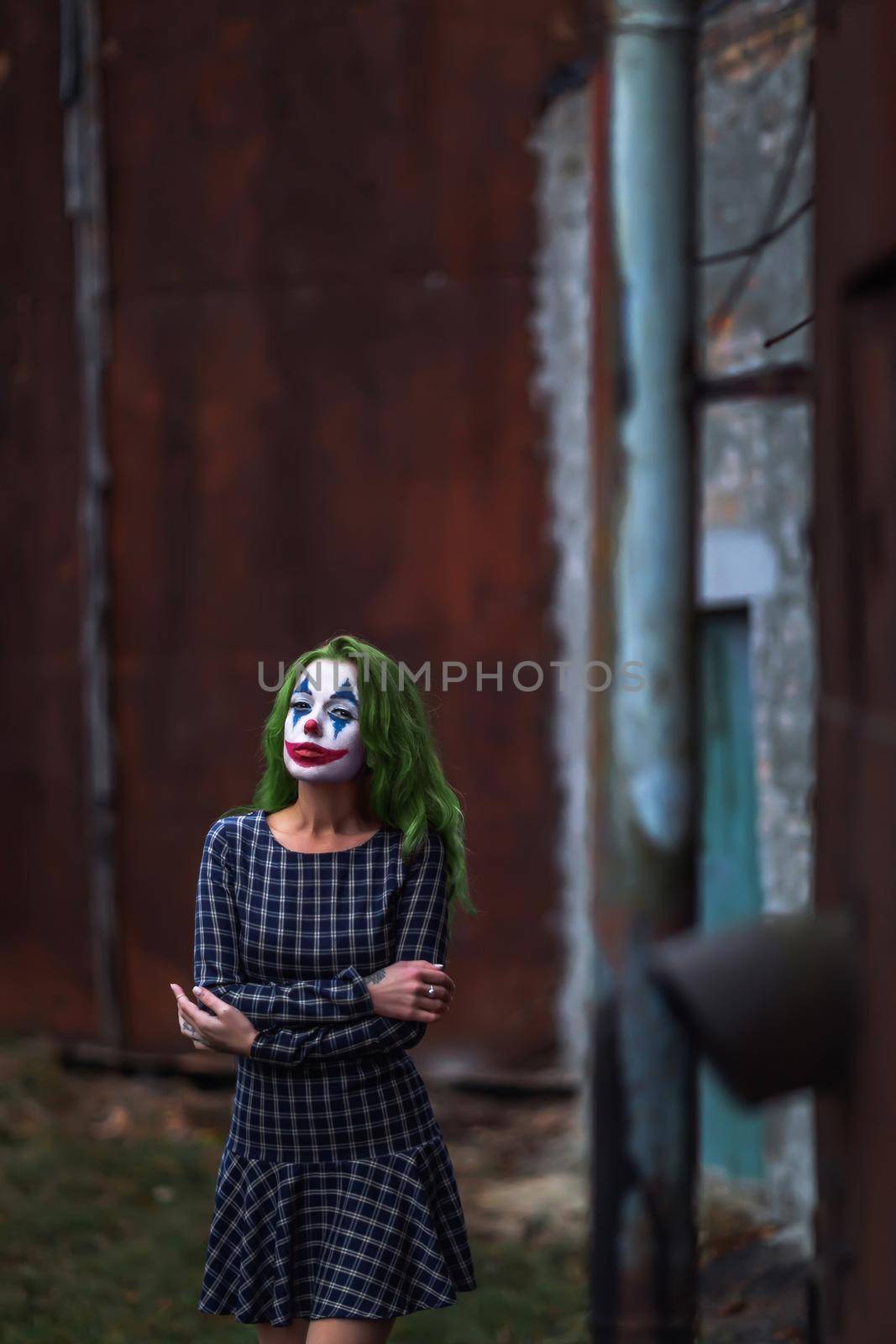 Portrait of a greenhaired girl in chekered dress with joker makeup on a atmospheric rusty metal wall background.