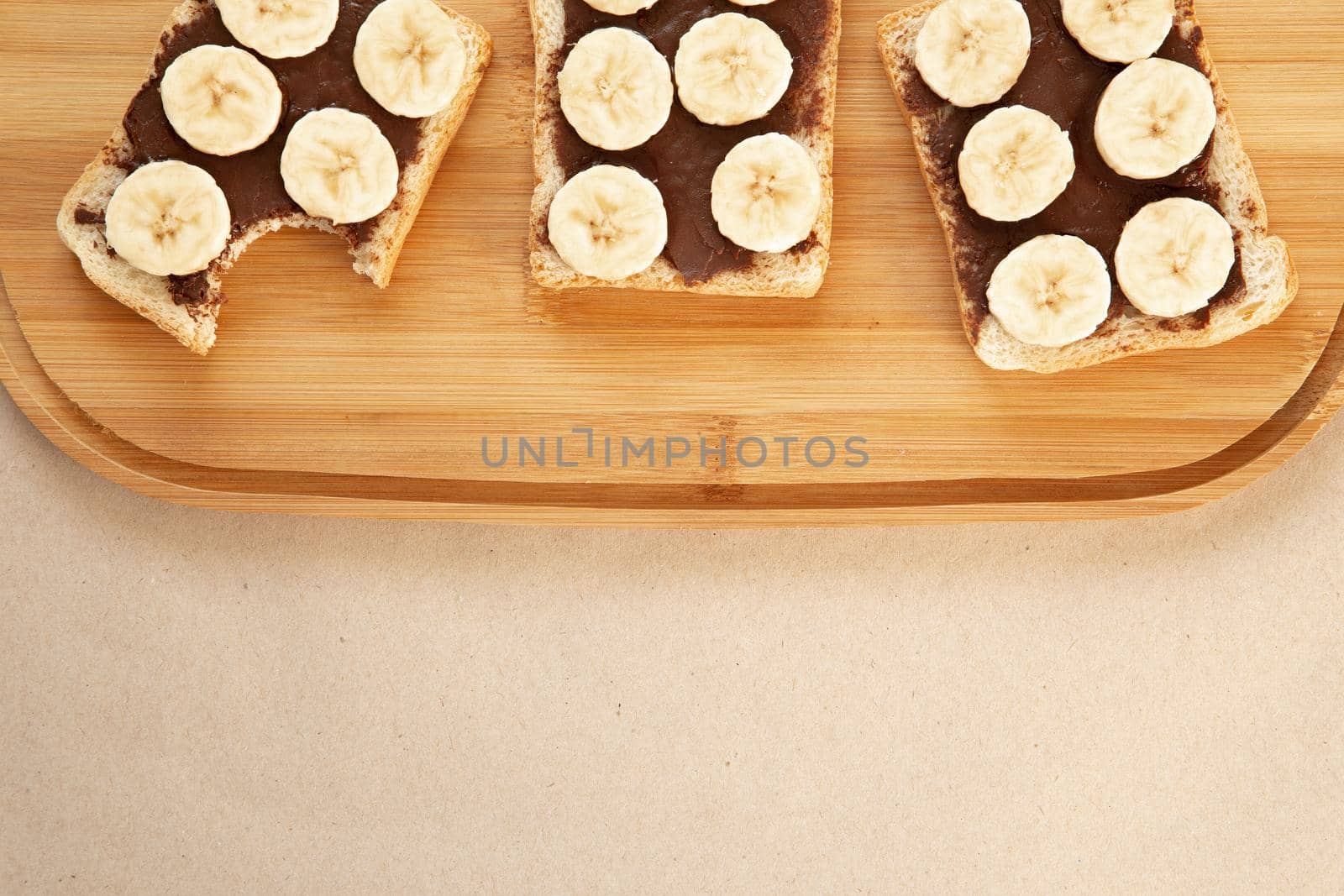 Three banana white bread toasts smeared with chocolate butter that lie on a cutting board on craft paper background. top view with area for text by lunarts