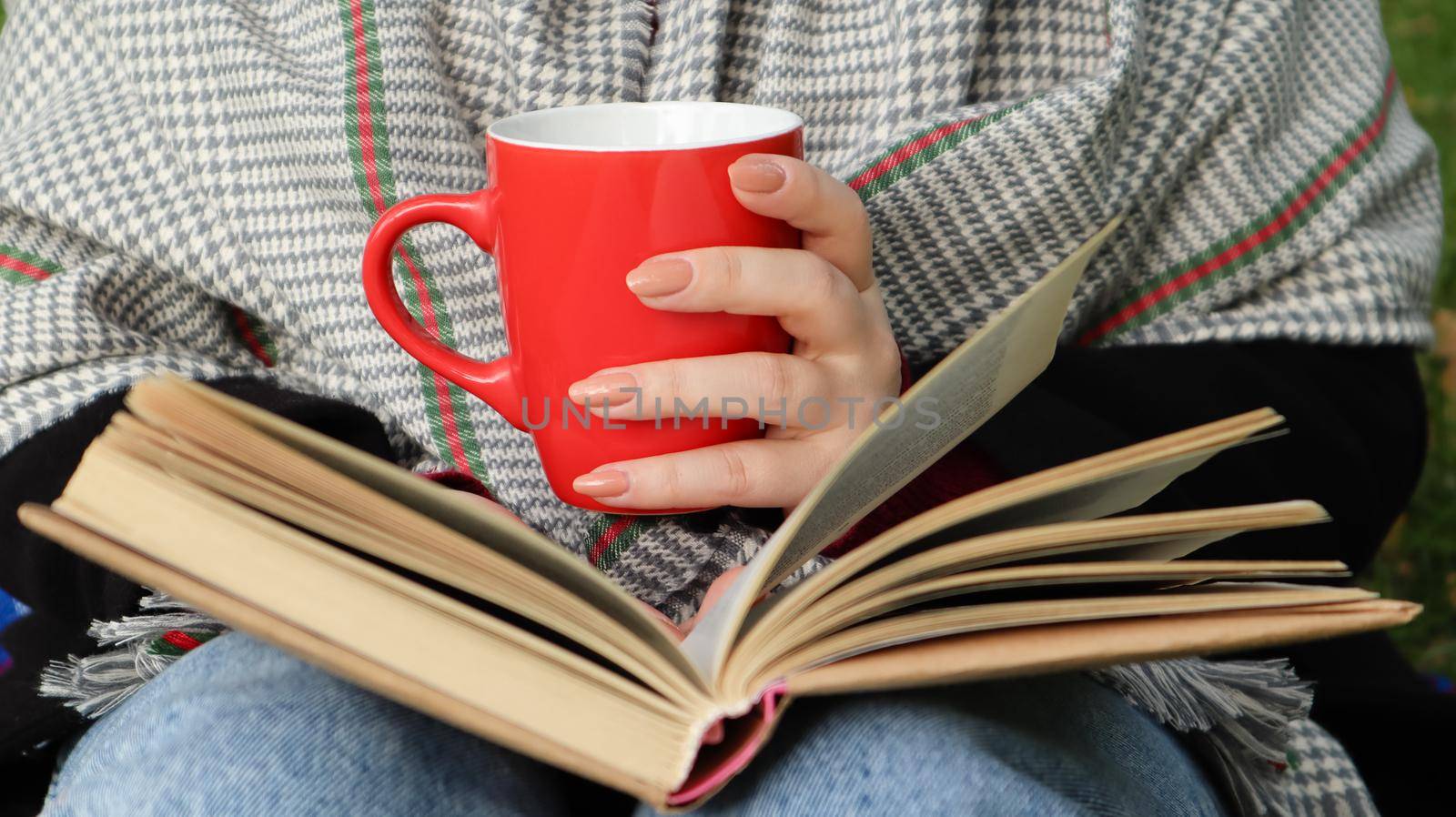 A girl dressed in a coat and a scarf in the autumn forest holds a book and a cup with a hot drink in her hands close-up in a city park on a warm day. The concept of reading, relaxation and comfort. by Roshchyn