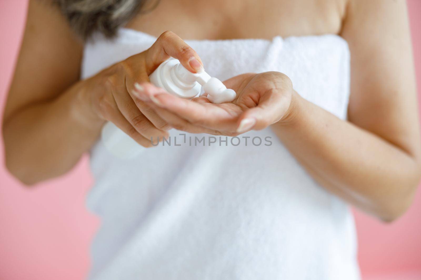 Middle aged woman with towel squeezes foam onto palm on pink background by Yaroslav_astakhov