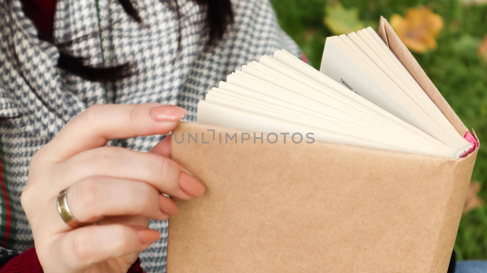 Girl reading a book in the autumn park. Female hands open the pages of a paper book outdoors on a warm sunny day. The student is preparing for the exam. Literary leisure in nature. by Roshchyn