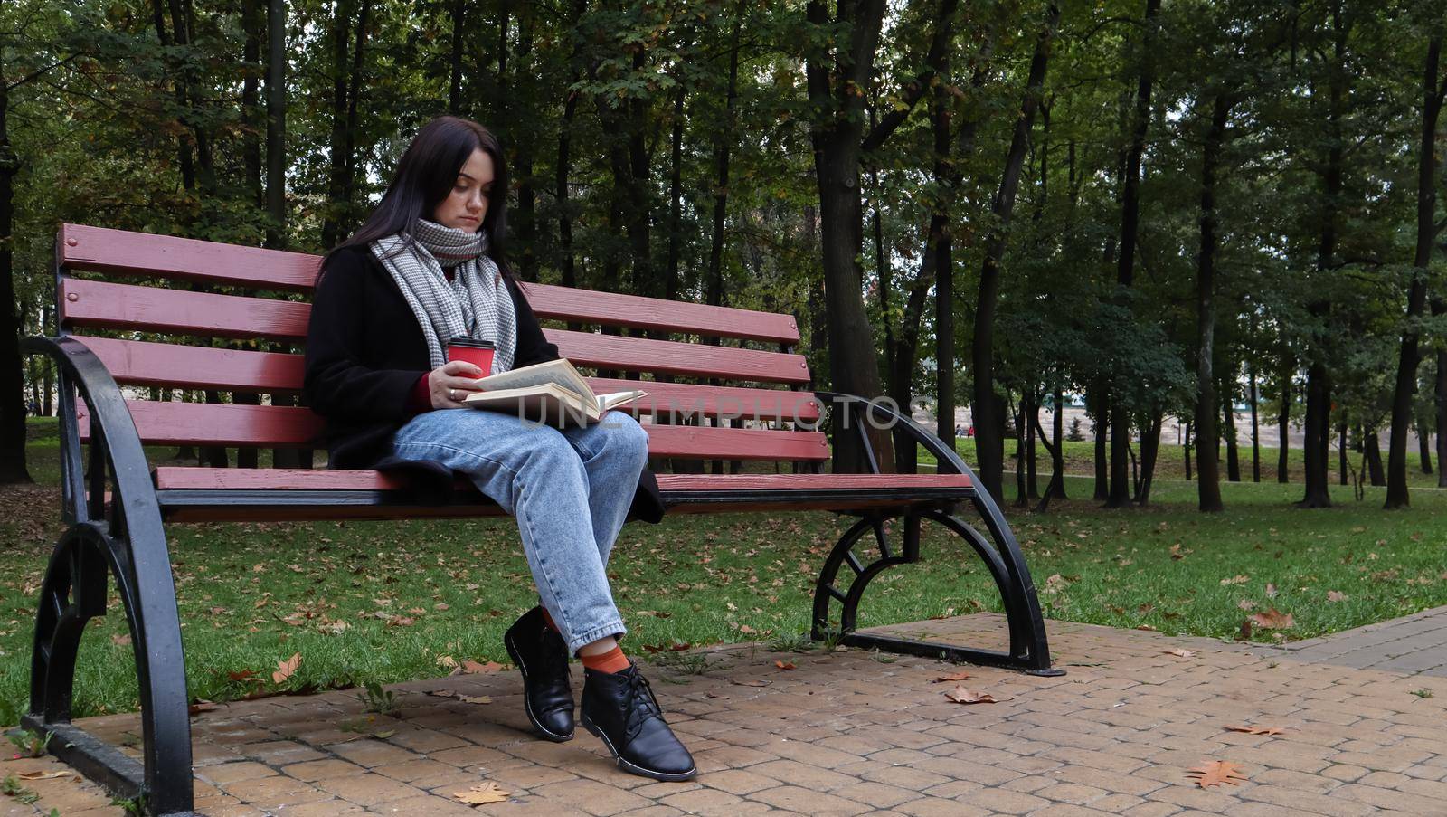 Young woman in jeans, coat and scarf, on a park bench. A woman is reading a book and drinking coffee or other hot drink outdoors alone. by Roshchyn