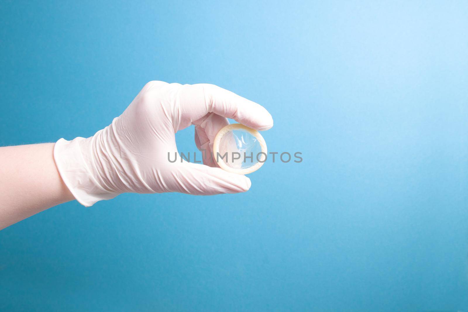 hand in a white medical rubber glove holds a condom without packaging on a blue background, copy space, close-up
