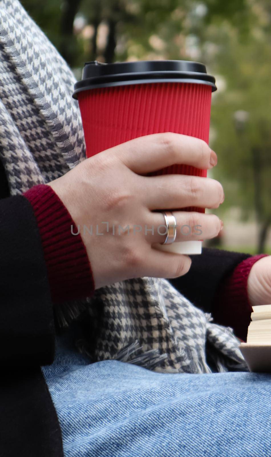 Young woman in jeans, coat and scarf, on a park bench. A woman is reading a book and drinking coffee or other hot drink outdoors alone. Close-up. The concept of honor, study, leisure and recreation. by Roshchyn