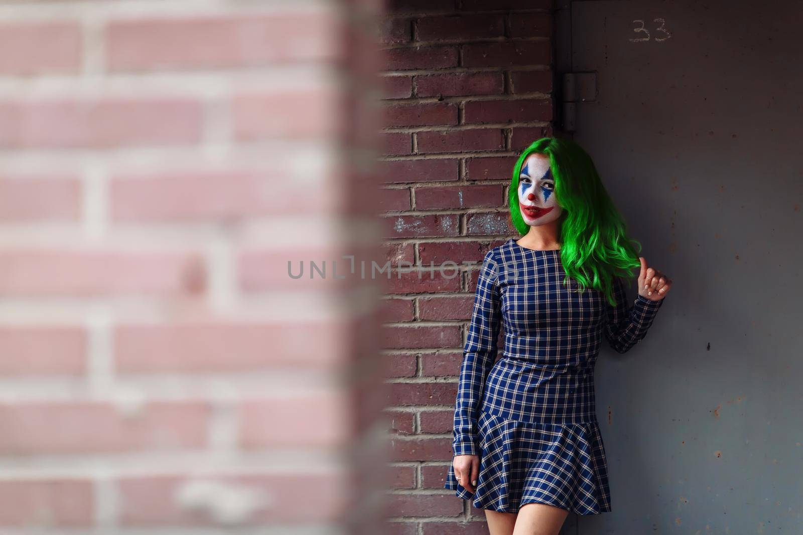Portrait of a greenhaired woman in chekered dress with joker makeup on a brick wall blurred background.