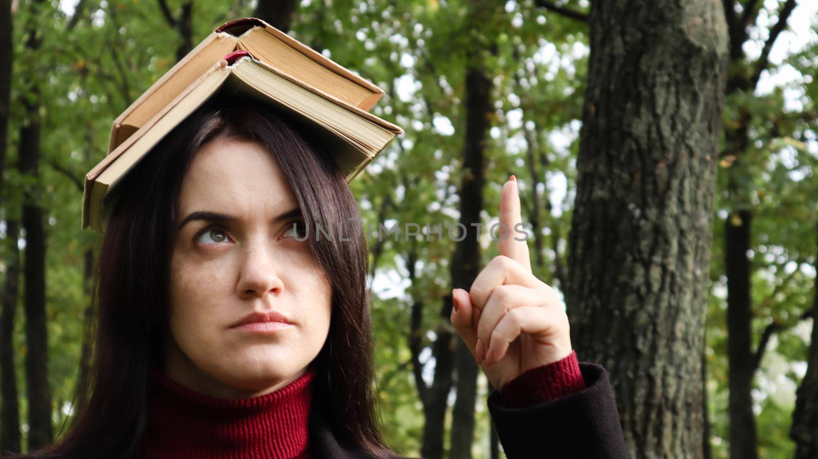 Portrait of a young and funny brunette in the park holding an open book on her head. Learning is fun. Woman balancing with books on her head. The student is tired of reading