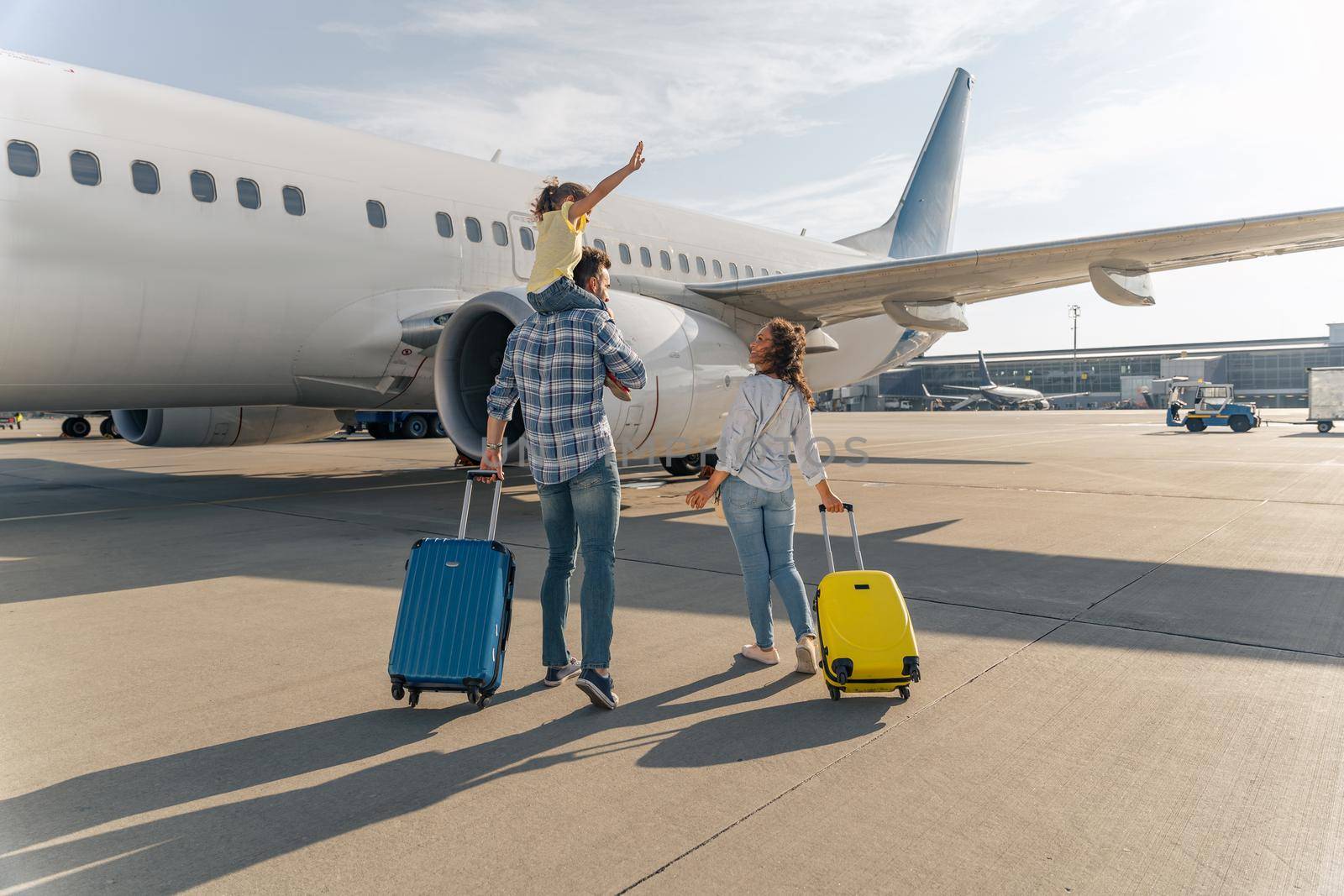 Back view of happy family standing near a large plane with two suitcases outdoor. Trip concept