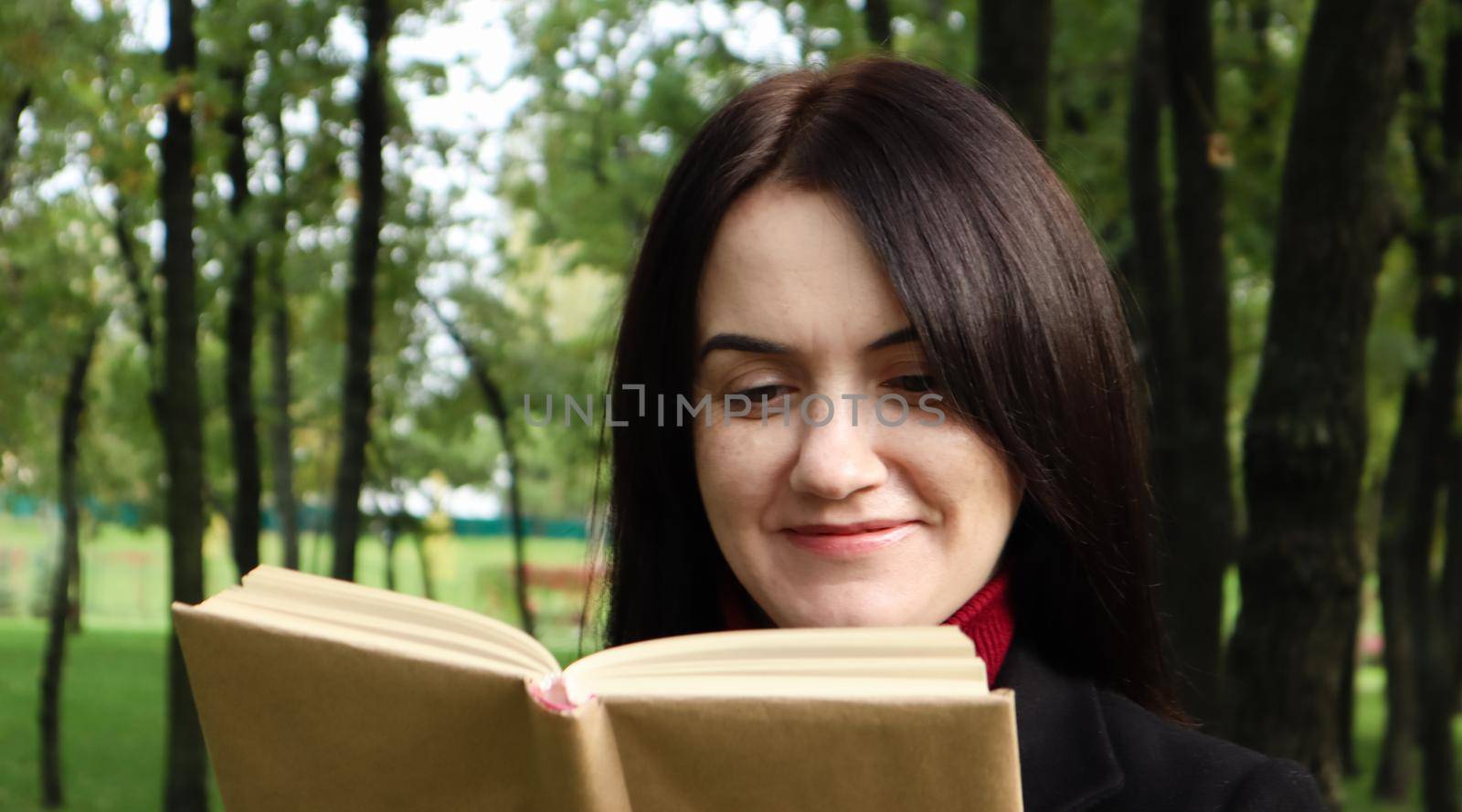 Smiling brunette woman reading a book in the park. Portrait of a happy charming Caucasian woman outdoors