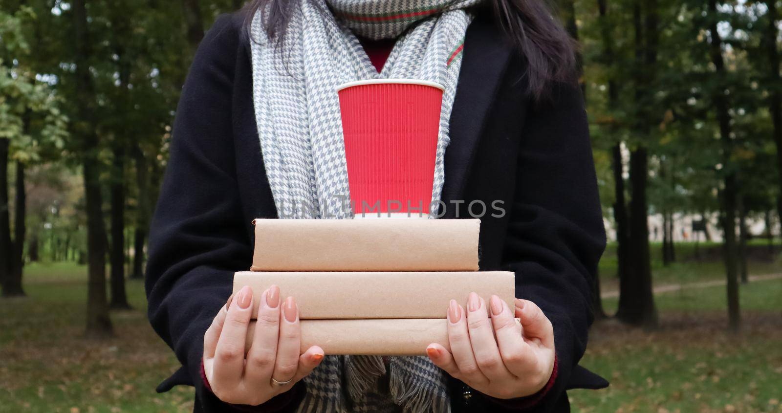 A young woman in a black coat and a gray scarf holds a stack of books and a glass with coffee in her hands on the background of an autumn park. On open air. Education, close hands of the girl