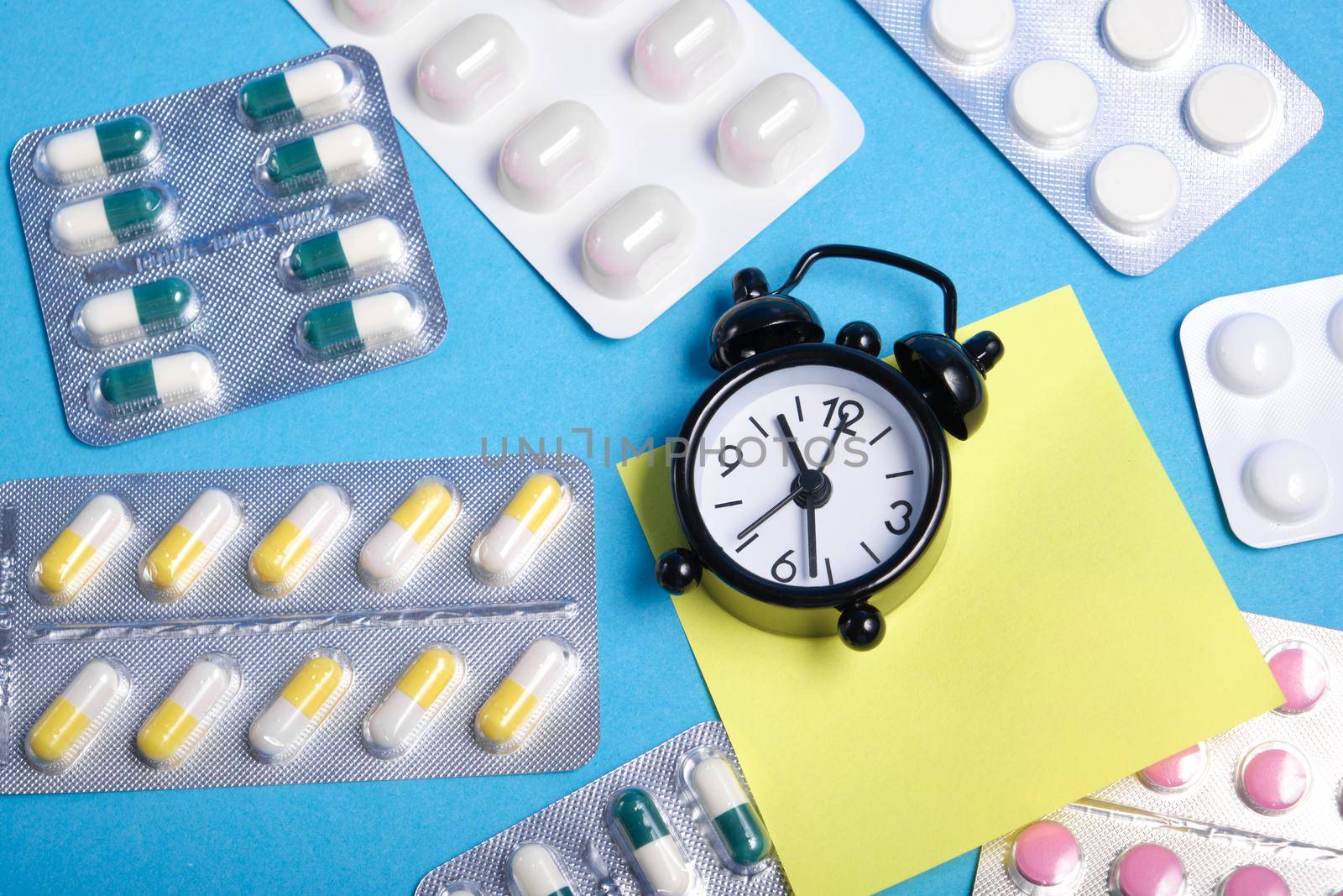 sticky note, black small alarm clock and packaging of pills on blue background, copy space, medication on time, medication intake reminder concept