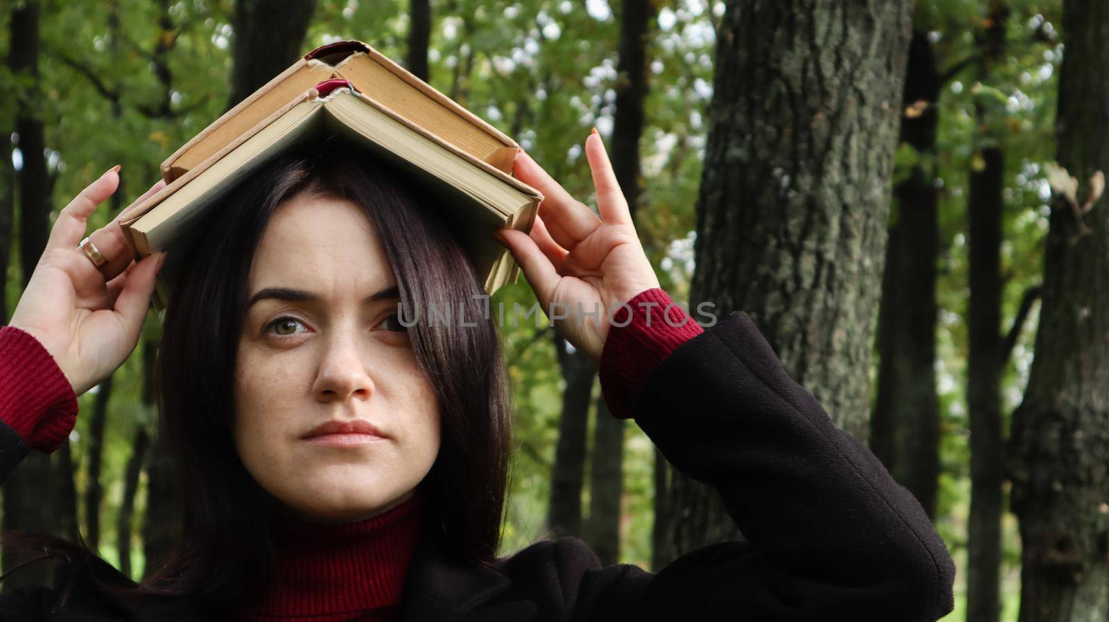 Portrait of a young and funny brunette in the park holding an open book on her head. Learning is fun. Woman balancing with books on her head. The student is tired of reading. by Roshchyn