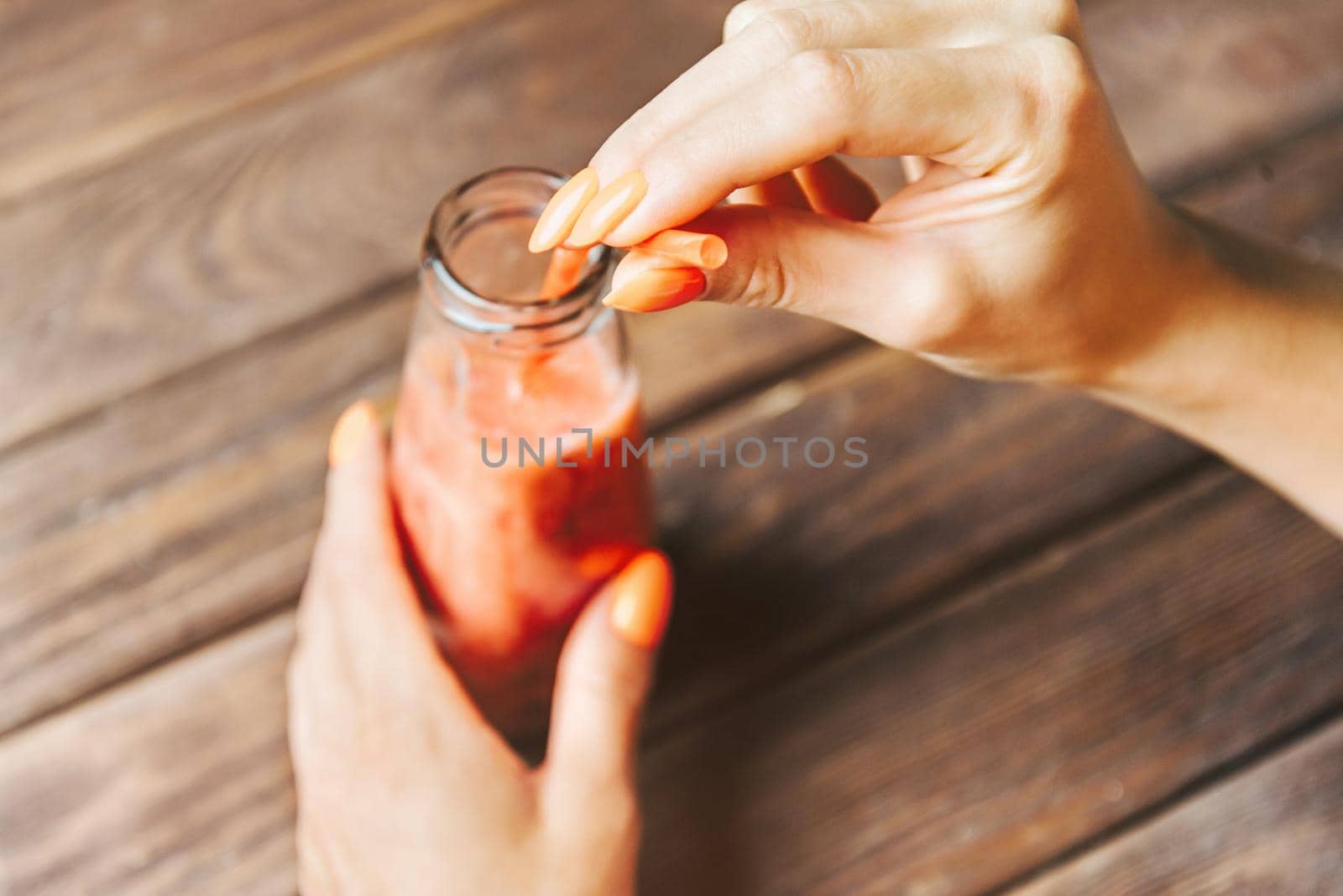 Young woman holding glass bottle of fresh smoothie drink with tube at wooden table. Point of view in first person.