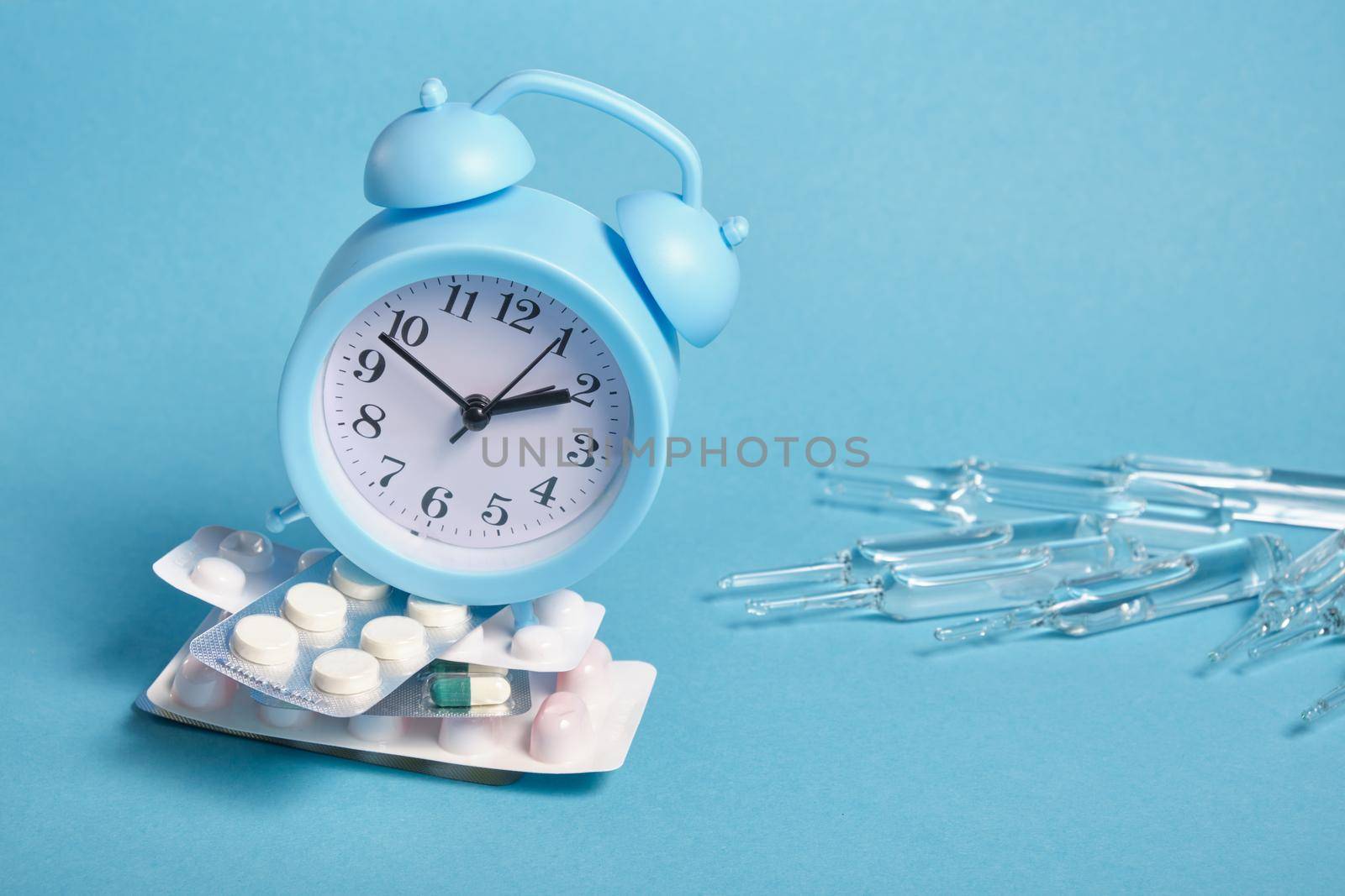large ampoules, light blue alarm clock on a pile of packaging of different pills on a blue background, copy space, taking medications on time concept