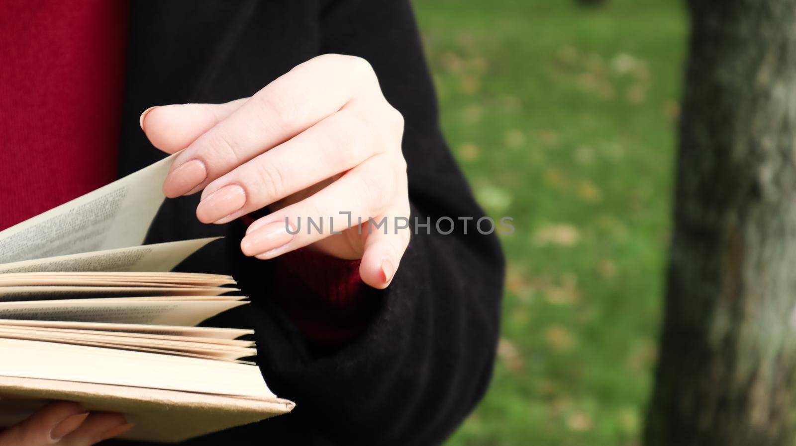 Girl reading a book in the park. Female hands flipping pages of paper book outdoors. The student is preparing for the exam. Literary leisure in nature. Close-up, copy space. by Roshchyn