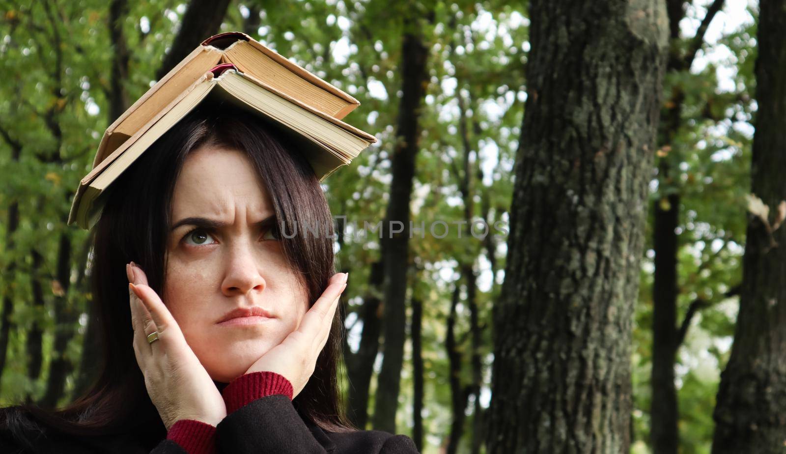 Portrait of a young and funny brunette in the park holding an open book on her head. Learning is fun. Woman balancing with books on her head. The student is tired of reading. by Roshchyn