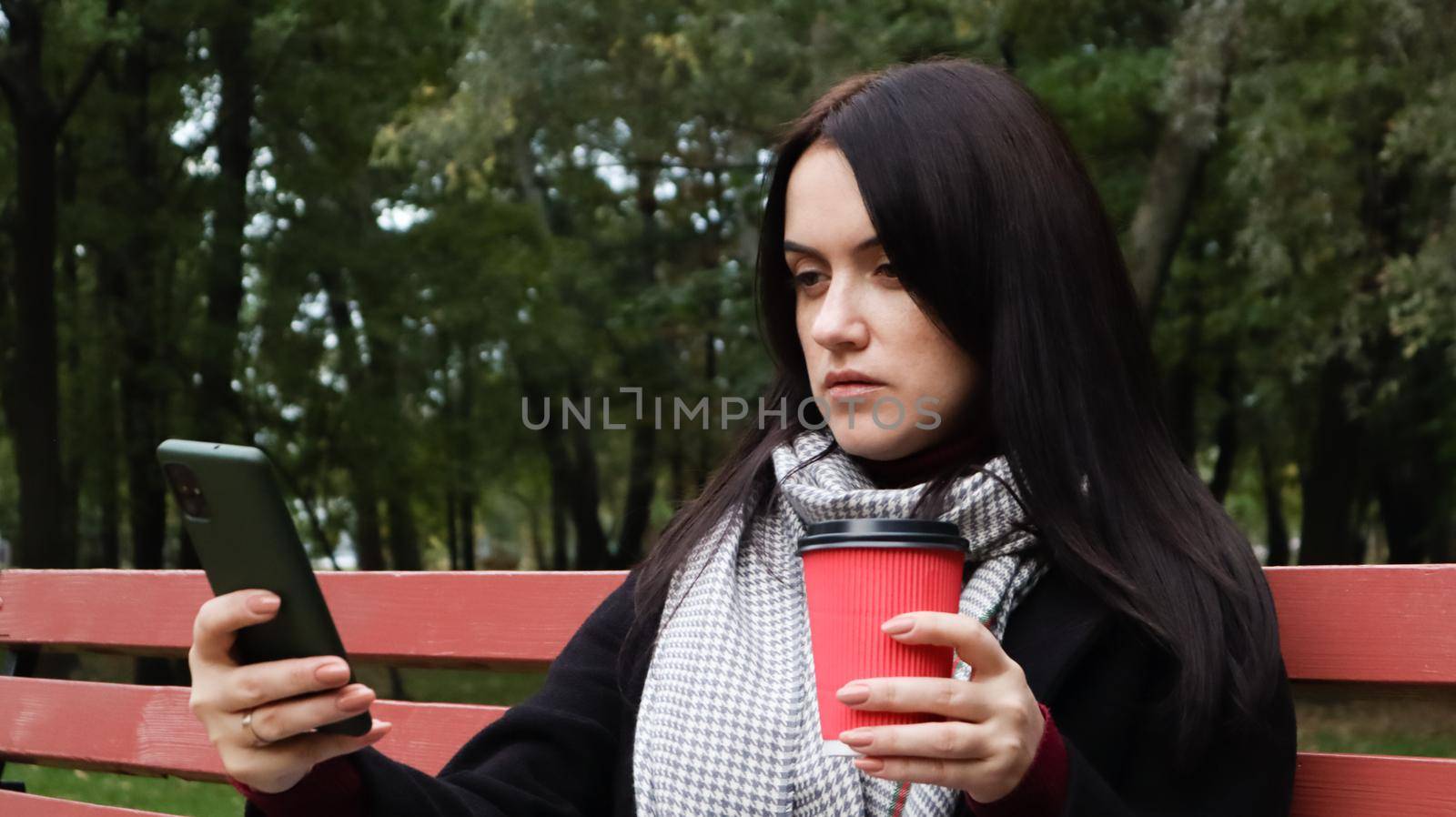 Beautiful young caucasian woman in a coat with a paper cup of takeaway coffee taking a selfie or taking pictures of herself for a blog with a smartphone outdoors in a park sitting on a bench