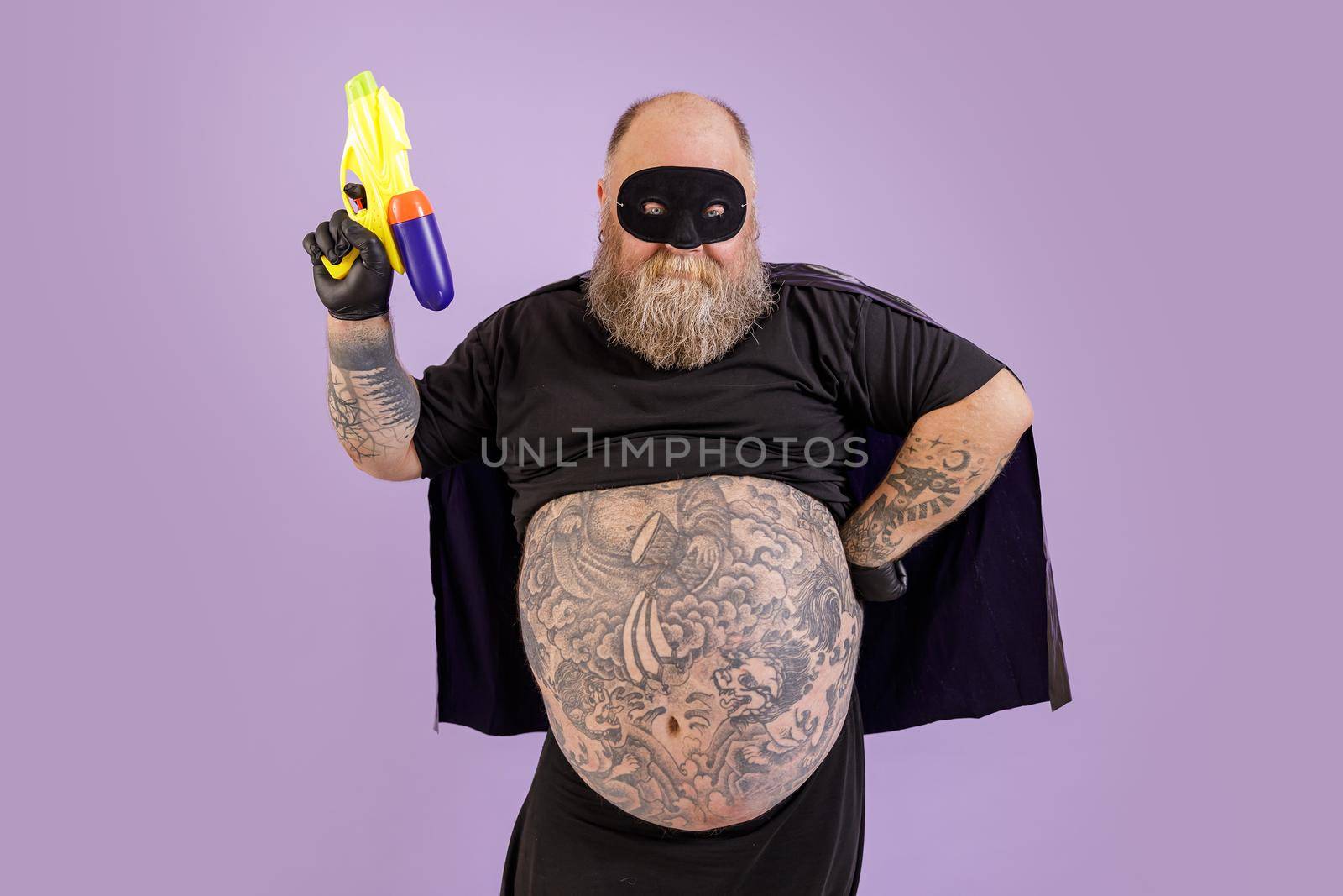 Mature bearded man with large tattooed abdomen wearing carnival costume with cape and mask holds toy blaster posing on purple background in studio