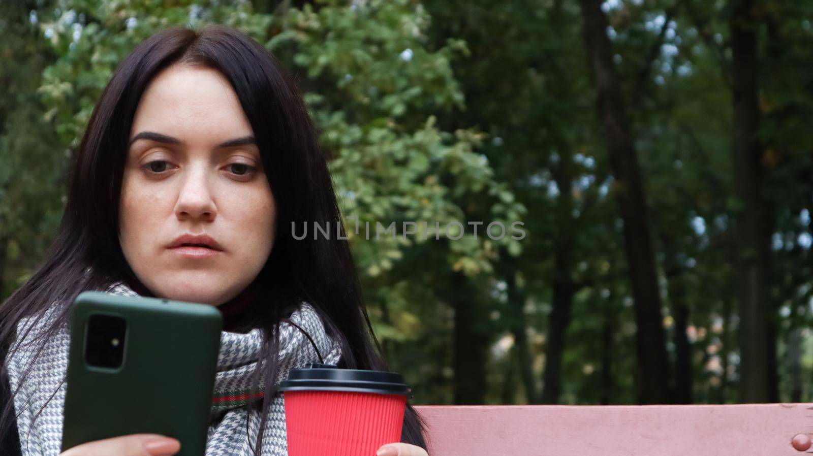 Beautiful young caucasian woman in a coat with a paper cup of takeaway coffee taking a selfie or taking pictures of herself for a blog with a smartphone outdoors in a park sitting on a bench. by Roshchyn