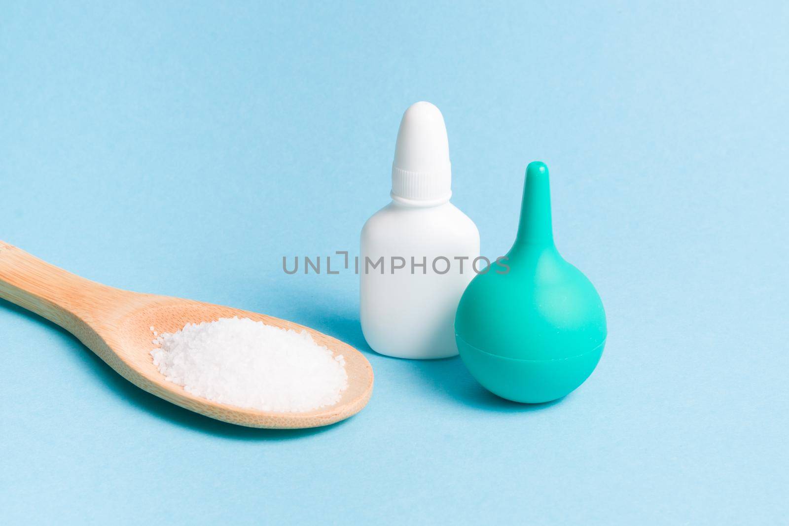 bottle of nose drops, a wooden spoon with sea food salt and a small green rubber enema for washing the nose on a light blue background