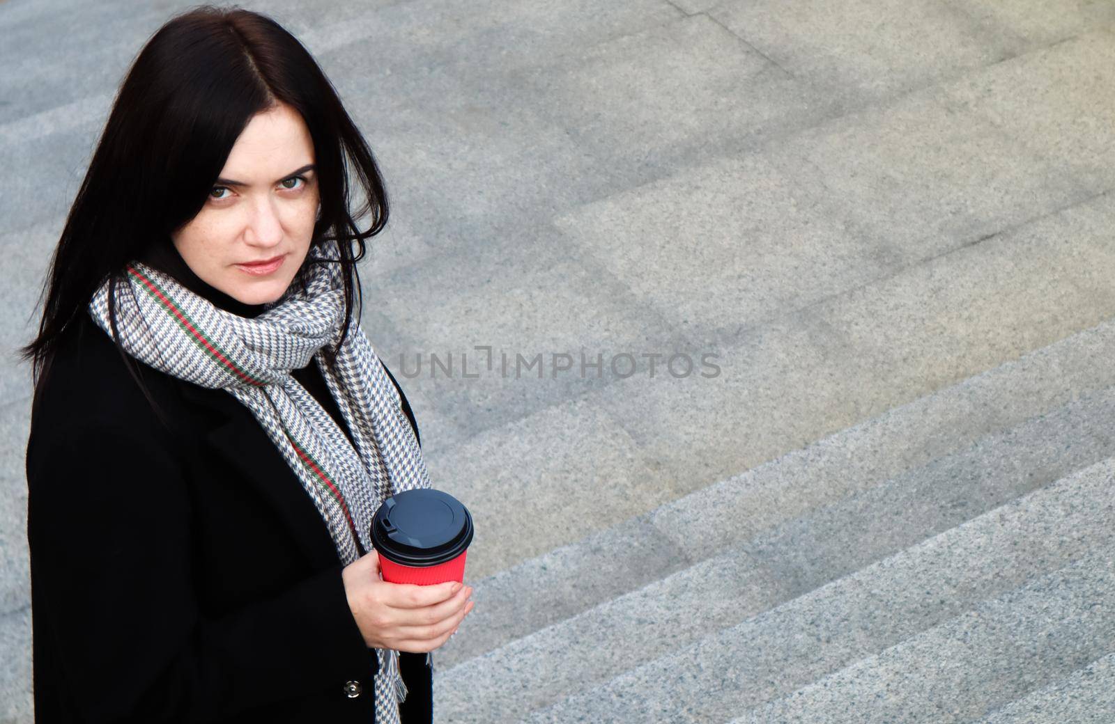 Young stylish woman in a coat and scarf drinks morning hot coffee in a red eco paper glass outdoors in an autumn park. Portrait of a young woman holding a takeaway coffee cup, shallow depth of field by Roshchyn