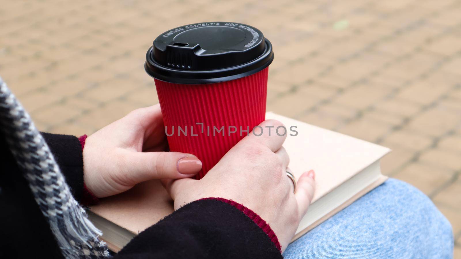 Young woman in jeans, coat and scarf, on a park bench. A woman is reading a book and drinking coffee or other hot drink outdoors alone. Close-up. The concept of honor, study, leisure and recreation