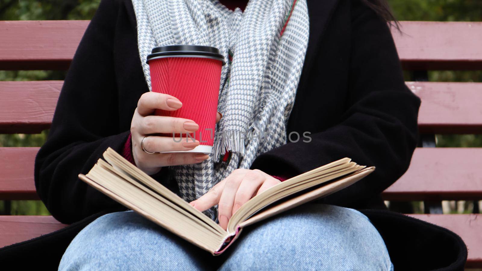 Young woman in jeans, coat and scarf, on a park bench. A woman is reading a book and drinking coffee or other hot drink outdoors alone. Close-up. The concept of honor, study, leisure and recreation. by Roshchyn