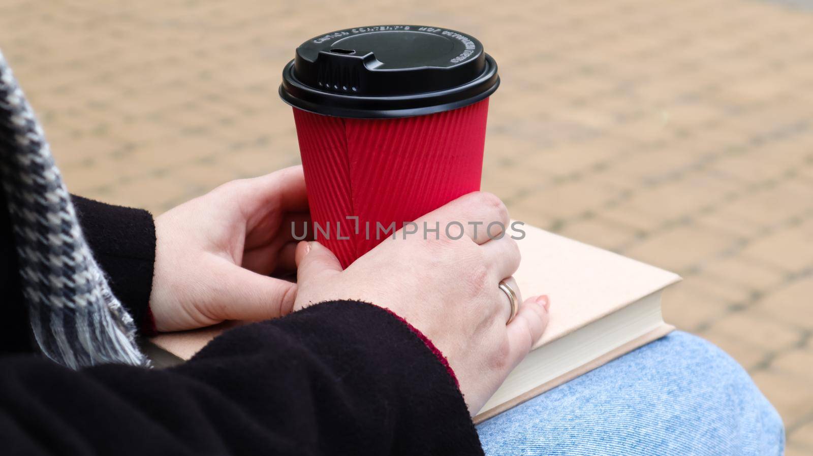 Young woman in jeans, coat and scarf, on a park bench. A woman is reading a book and drinking coffee or other hot drink outdoors alone. Close-up. The concept of honor, study, leisure and recreation