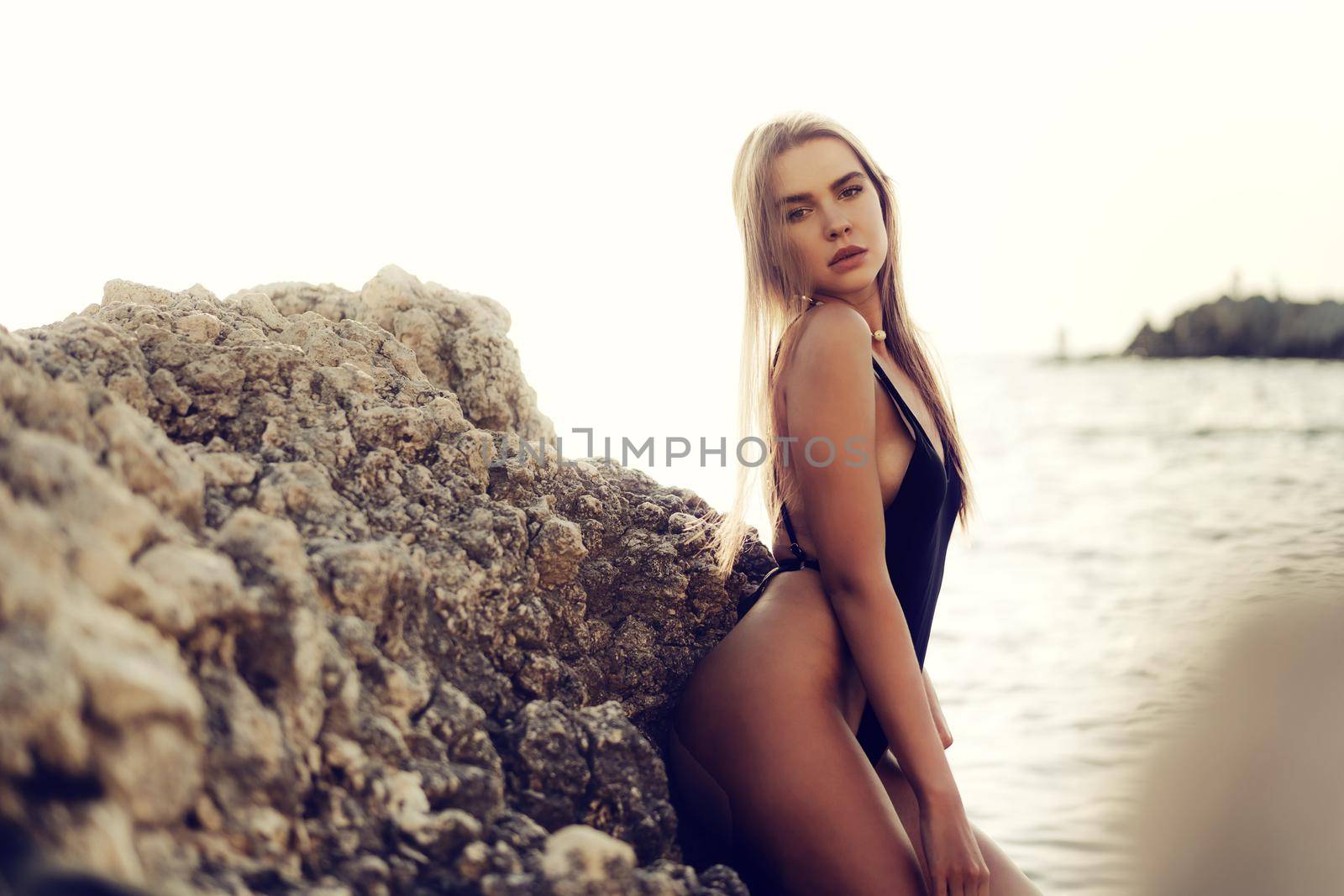 Young beautiful sexy female model in black bikini standing in sea water near huge rock