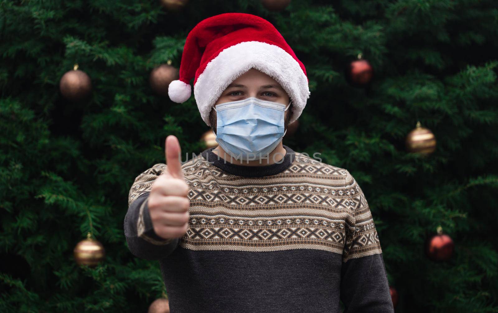 Christmas thumbs up like sign. Close up Portrait of man wearing a santa claus hat, xmas sweater and medical mask with emotion. Against the background of a Christmas tree. Coronavirus pandemic