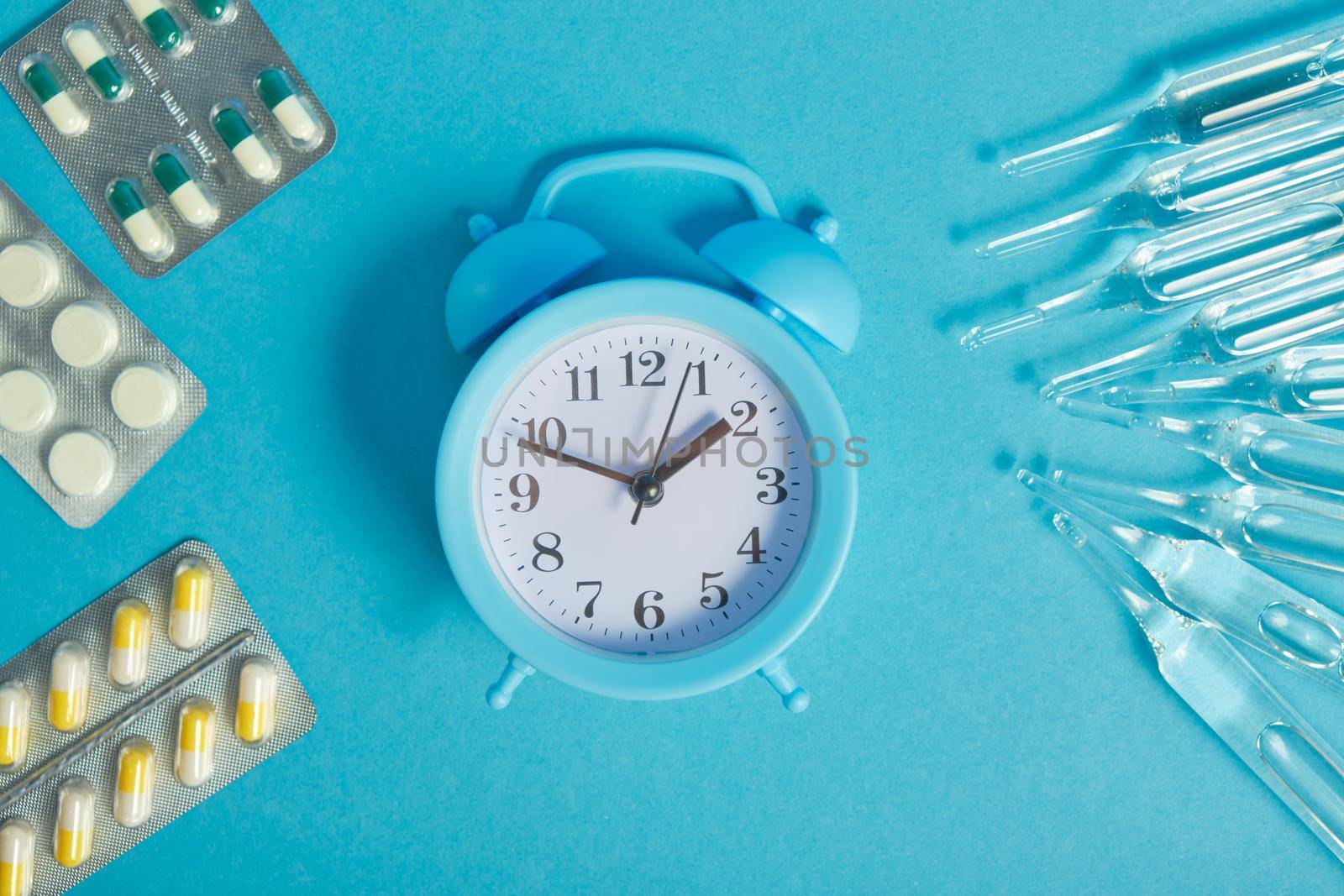 light blue alarm clock and medicines on a blue background, ampoules and packs of pills around the clock, time to take medicines
