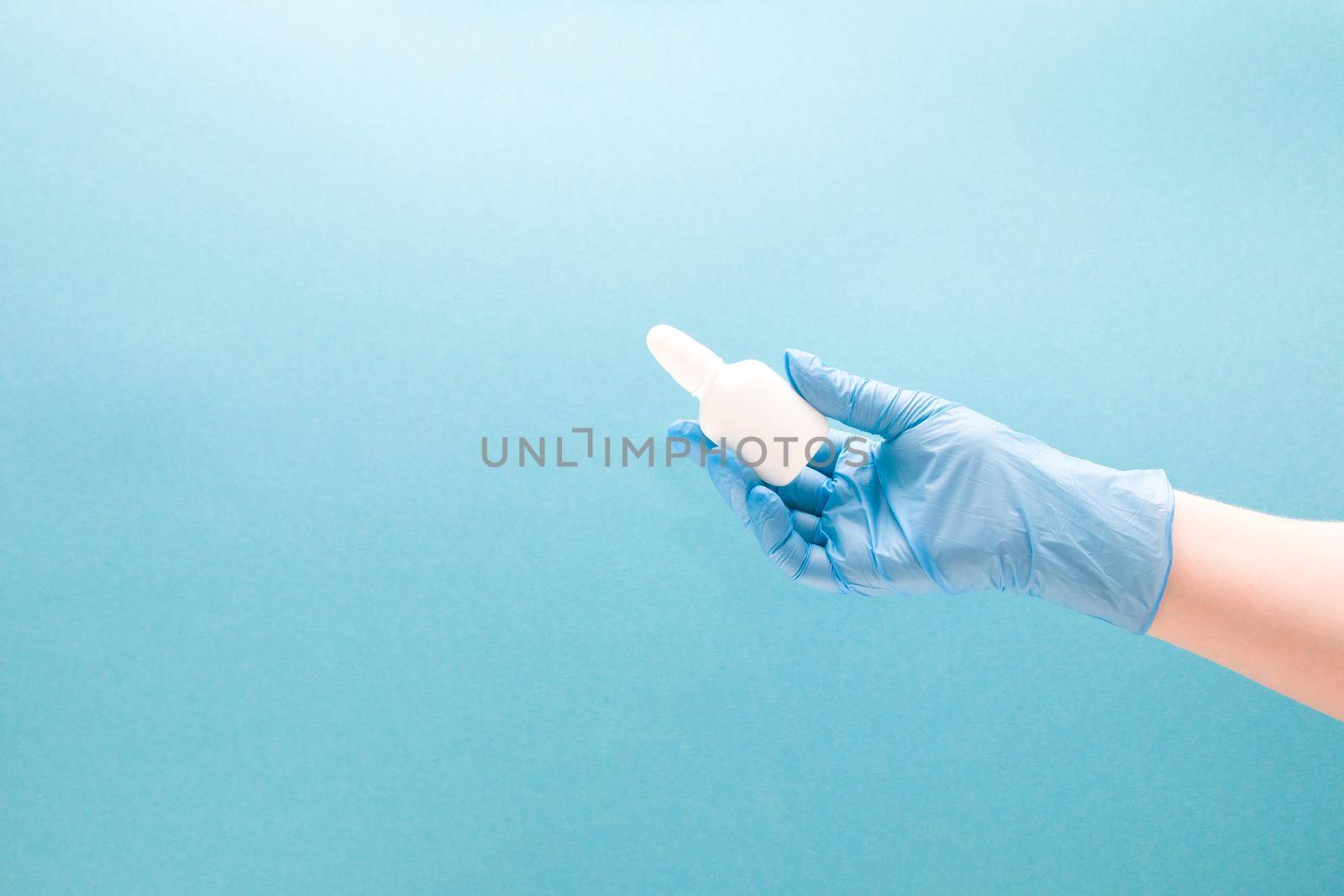 a female hand in a blue disposable medical glove holds a white plastic bottle with drops for the nose, blue background copy space, nasal drops, medicine for the nose