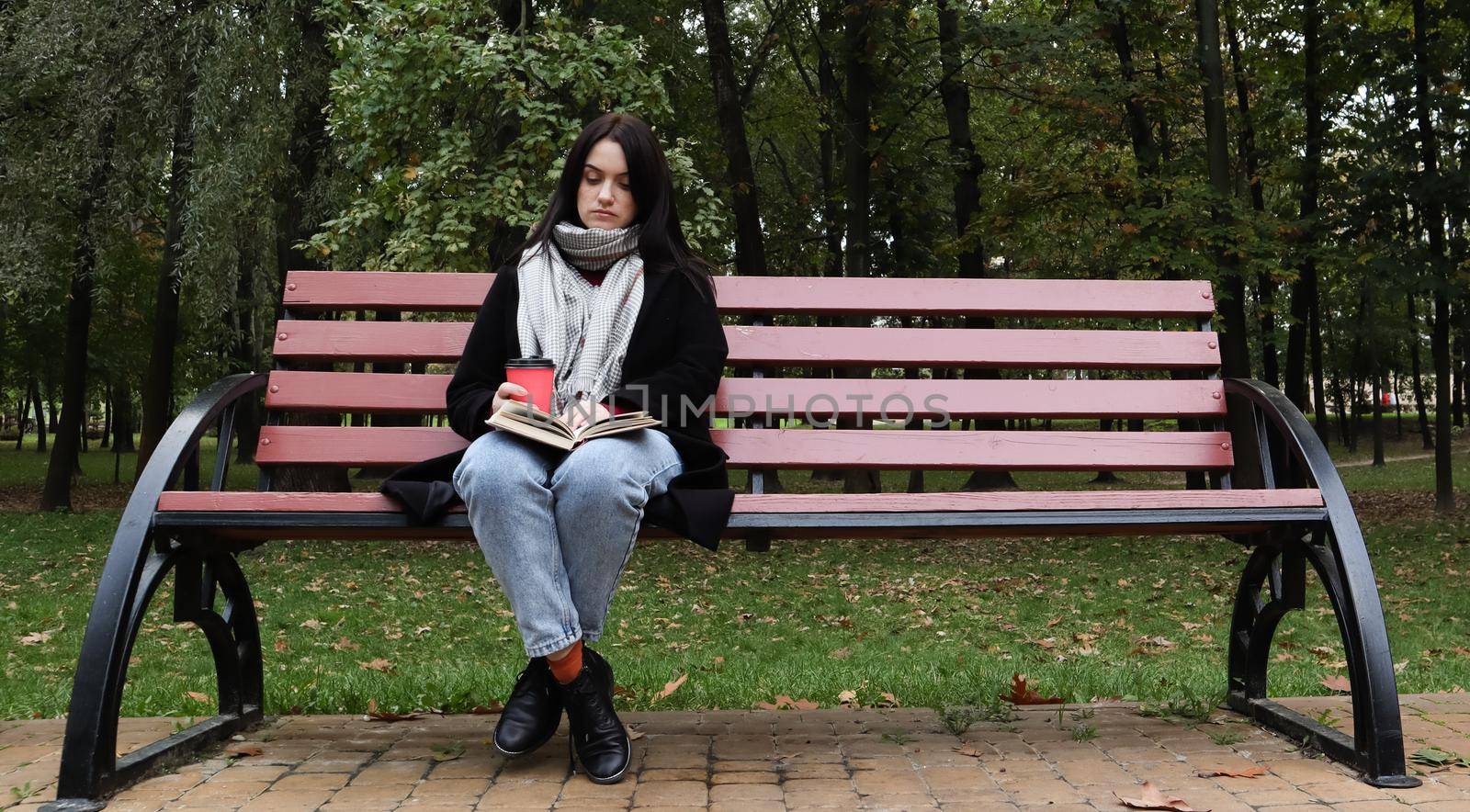 Young woman in jeans, coat and scarf, on a park bench. A woman is reading a book and drinking coffee or other hot drink outdoors alone