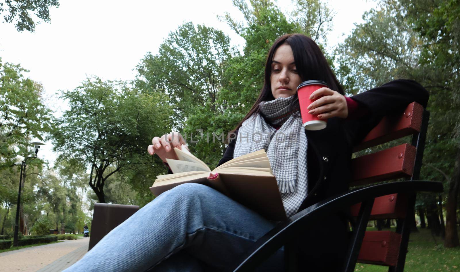 Young woman in jeans, coat and scarf, on a park bench. A woman is reading a book and drinking coffee or other hot drink outdoors alone. by Roshchyn