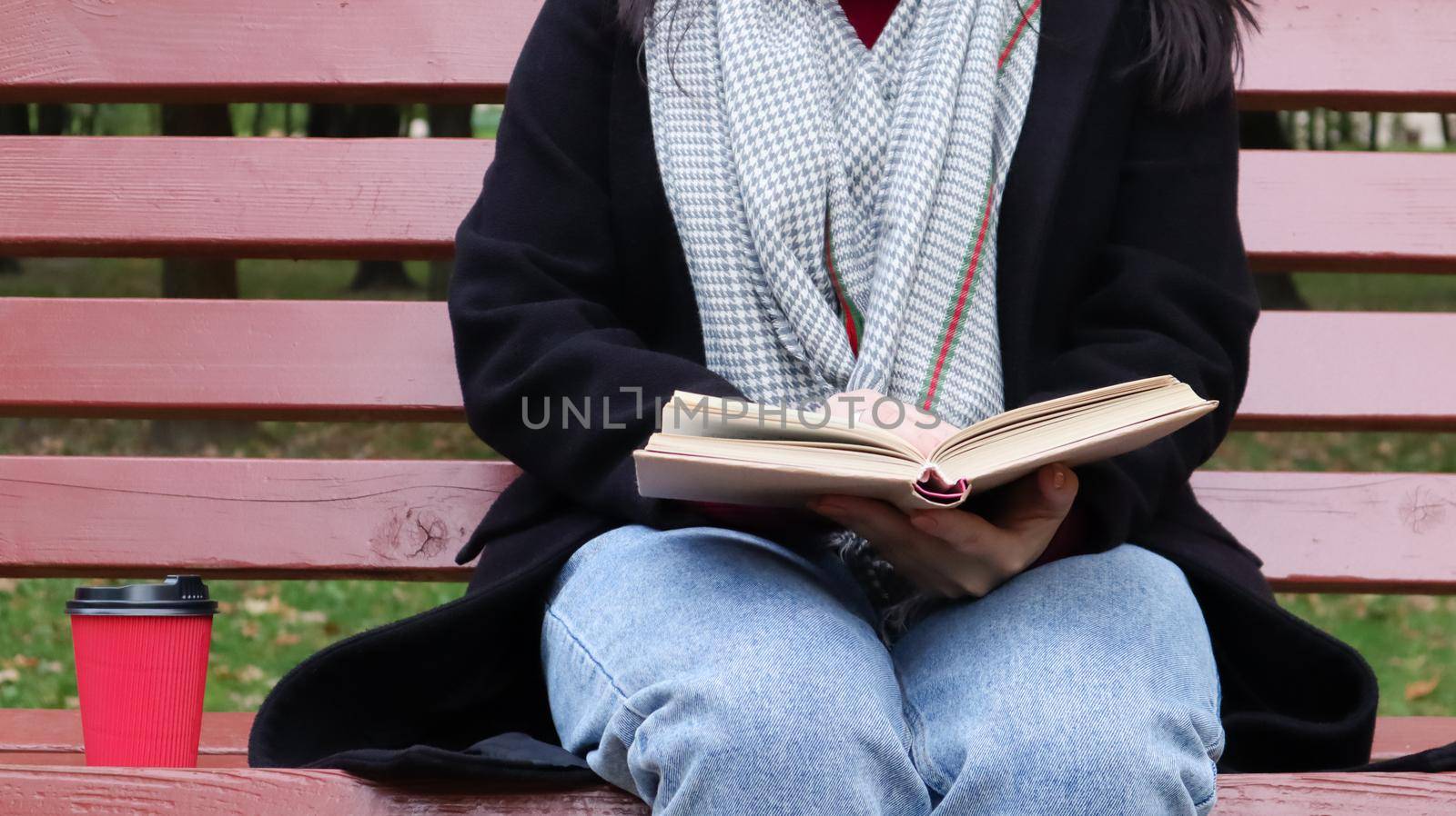 Young woman in jeans, coat and scarf, on a park bench. A woman is reading a book and drinking coffee or other hot drink outdoors alone. Close-up. The concept of honor, study, leisure and recreation. by Roshchyn