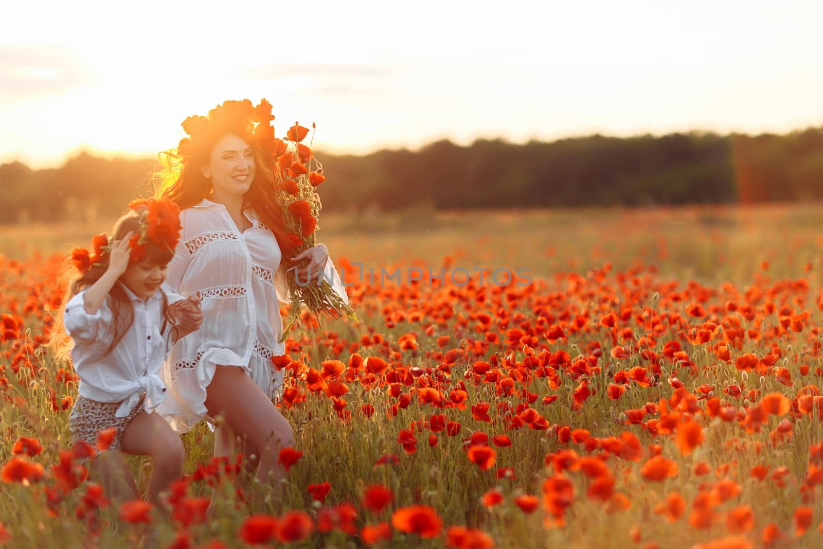 Little girl with redhead mother in white dresses and wreathes walking with bouquet of poppies on poppy field at warm summer sunset