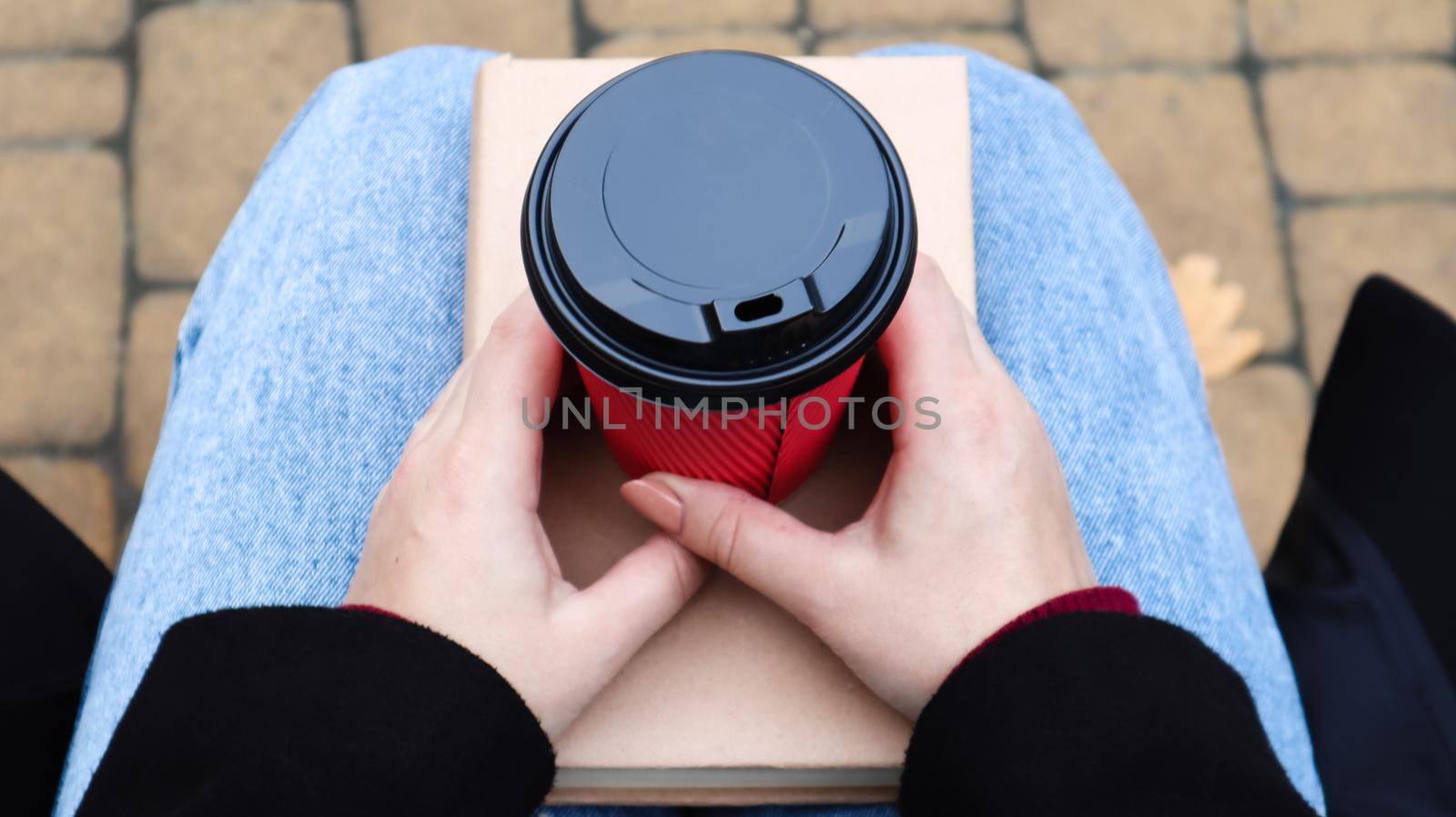 Young woman in jeans, coat and scarf, on a park bench. A woman is reading a book and drinking coffee or other hot drink outdoors alone. Close-up. View from above. The concept of honor, leisure