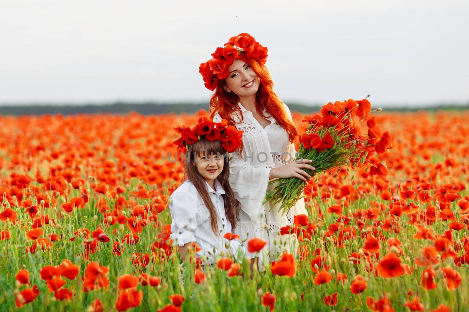 Little girl with redhead mother in white dresses and wreathes poses with bouquet of poppies on poppy field at summer sunset by OnPhotoUa