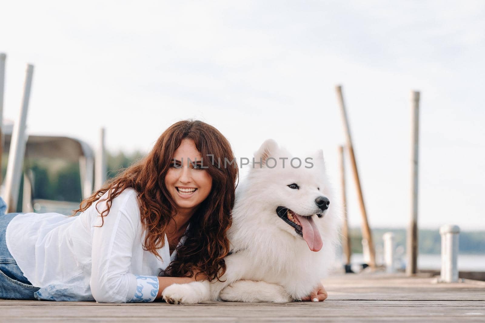 a happy woman with a big white dog lies on a pier near the sea at sunset.
