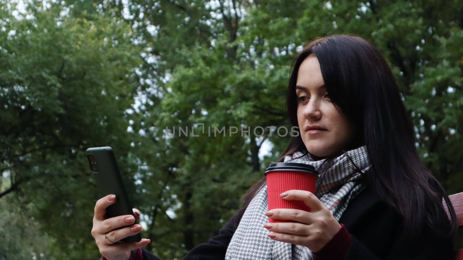 Beautiful young caucasian woman in a coat with a paper cup of takeaway coffee taking a selfie or taking pictures of herself for a blog with a smartphone outdoors in a park sitting on a bench. by Roshchyn