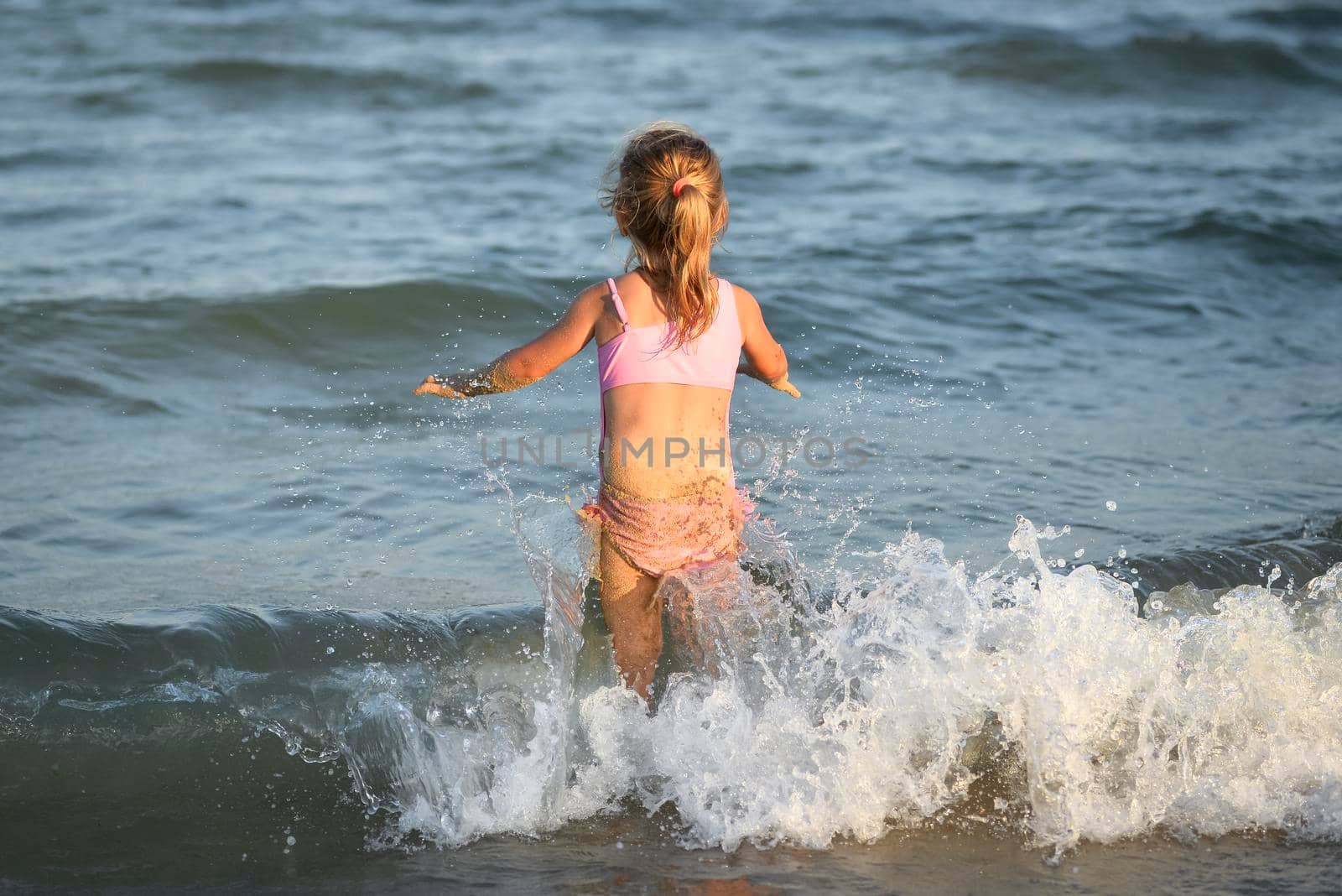 Girl in pink swimsuit standing on the beach and looking at the morning sea with waves. by esvetleishaya