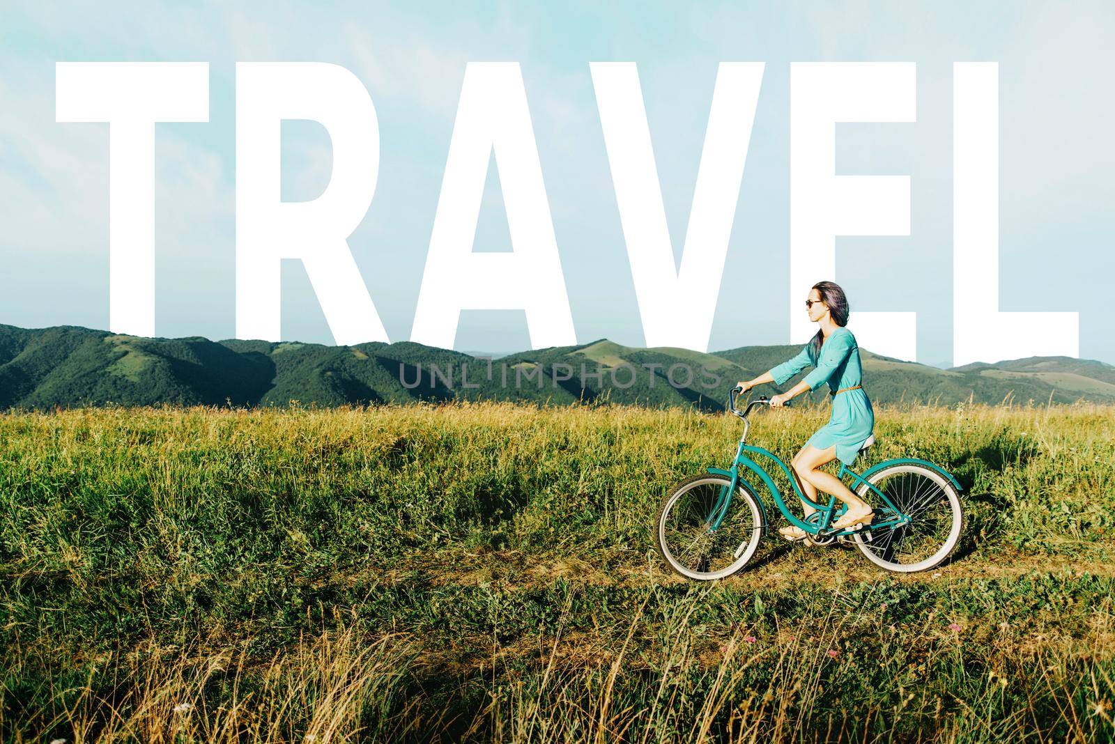 Happy beautiful young woman in dress riding a bicycle on summer meadow on background of word travel.