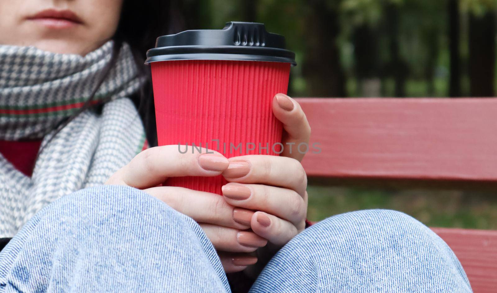 Young stylish woman in a coat and scarf drinks morning hot coffee in a red eco paper glass outdoors in an autumn park. Close-up of a young woman holding a takeaway coffee cup, shallow depth of field.