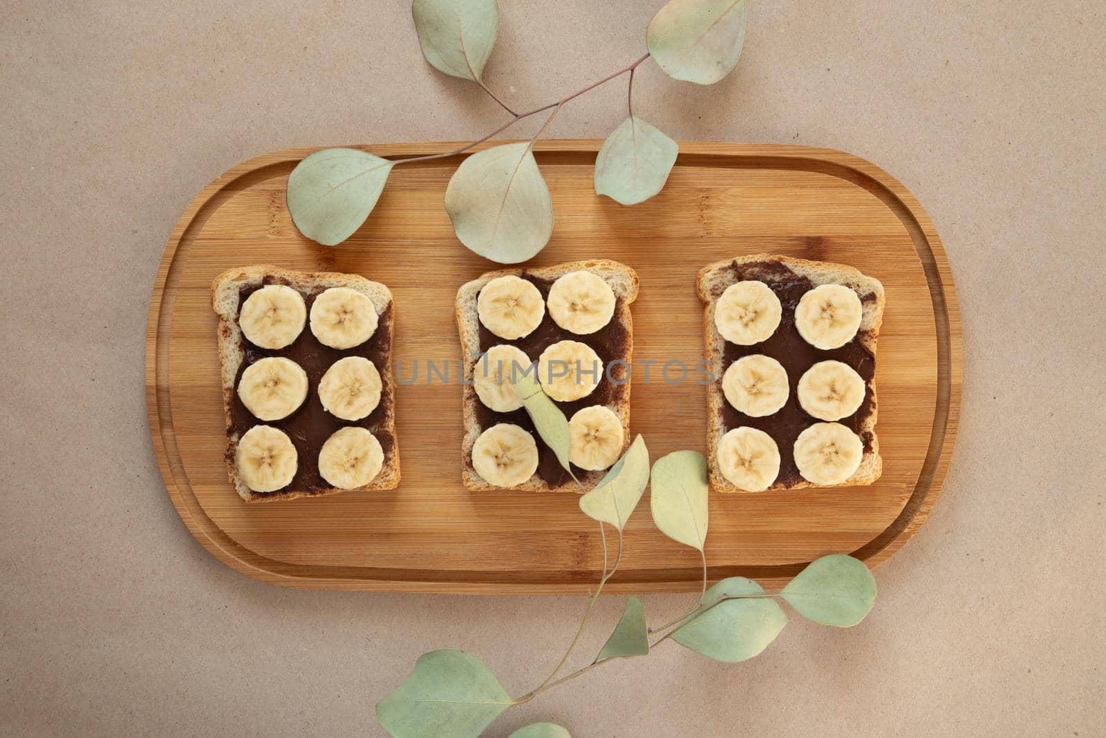 Three banana white bread toasts smeared with chocolate butter that lie on a cutting board with a sprig of leaves on craft paper background. top view with area for text by lunarts