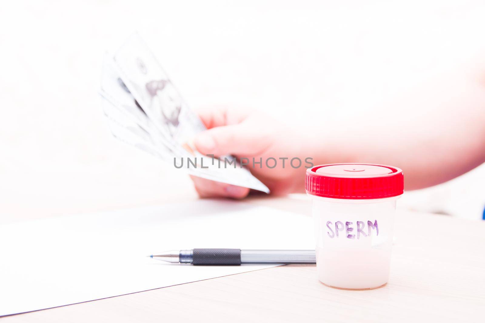 test jar with a red lid and the inscription sperm, against the background a man holds several notes of 100 dallors, a copy space, sperm in a jar, a sperm donor concept, sell sperm, a pen and paper on the table