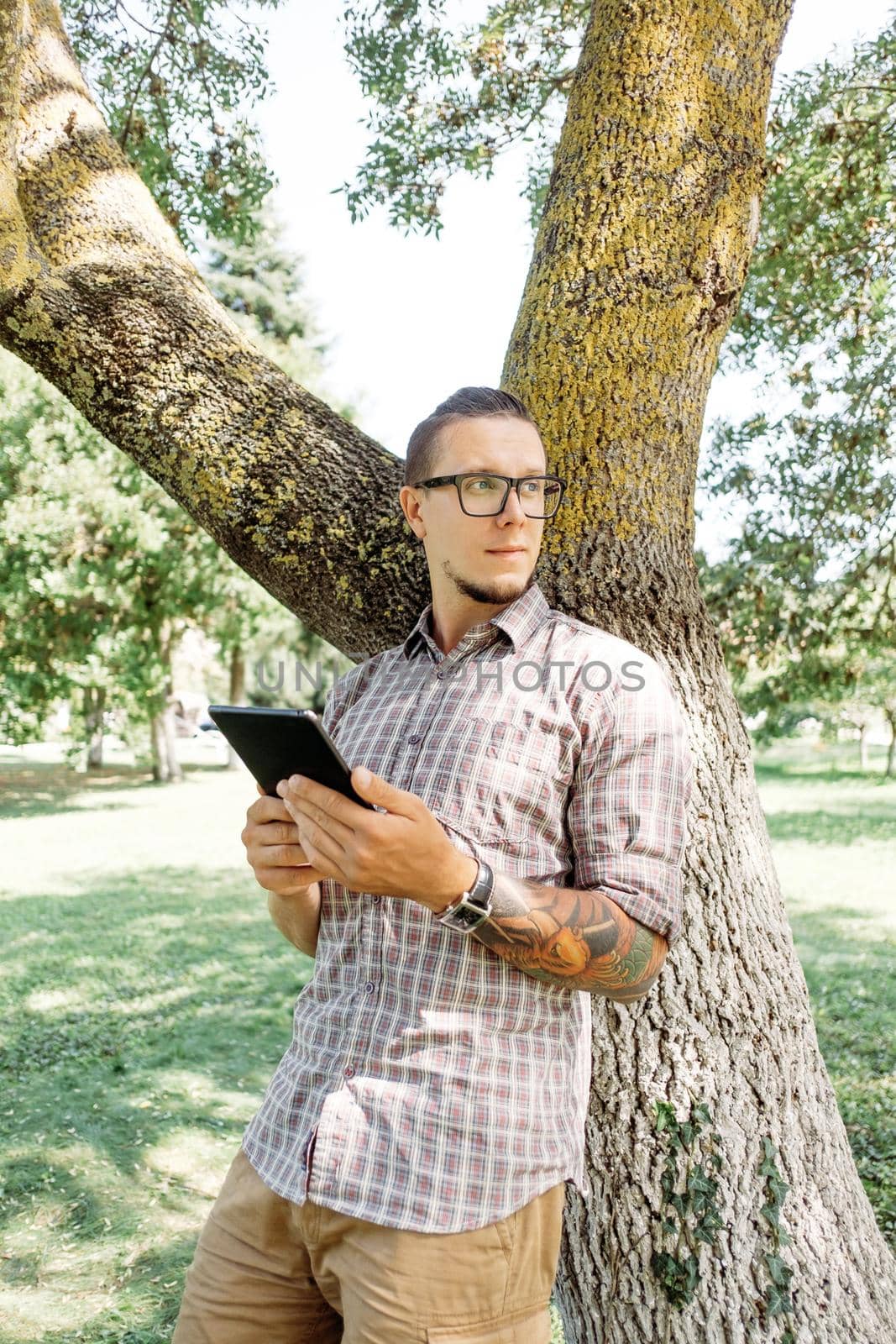 Handsome casual style young man standing with digital tablet near the tree in park.