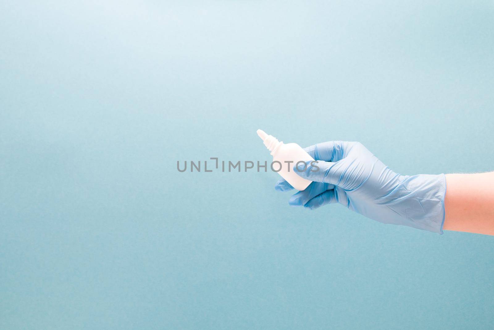 a female hand in a blue disposable medical glove holds a white plastic bottle with drops for the nose without a cap, blue background copy space, nasal drops, medicine for the nose by natashko