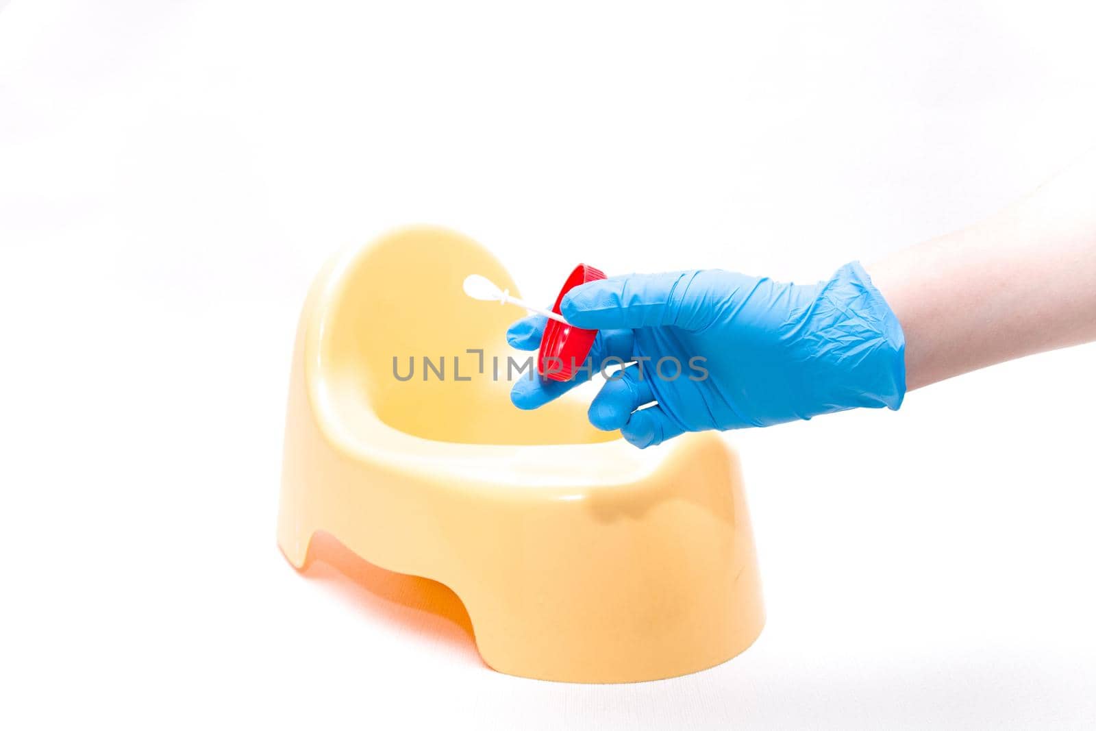 a hand in a disposable blue medical glove holds a test jar with a white spoon for collecting feces, in the background is a yellow children's pot, white background, copy space by natashko